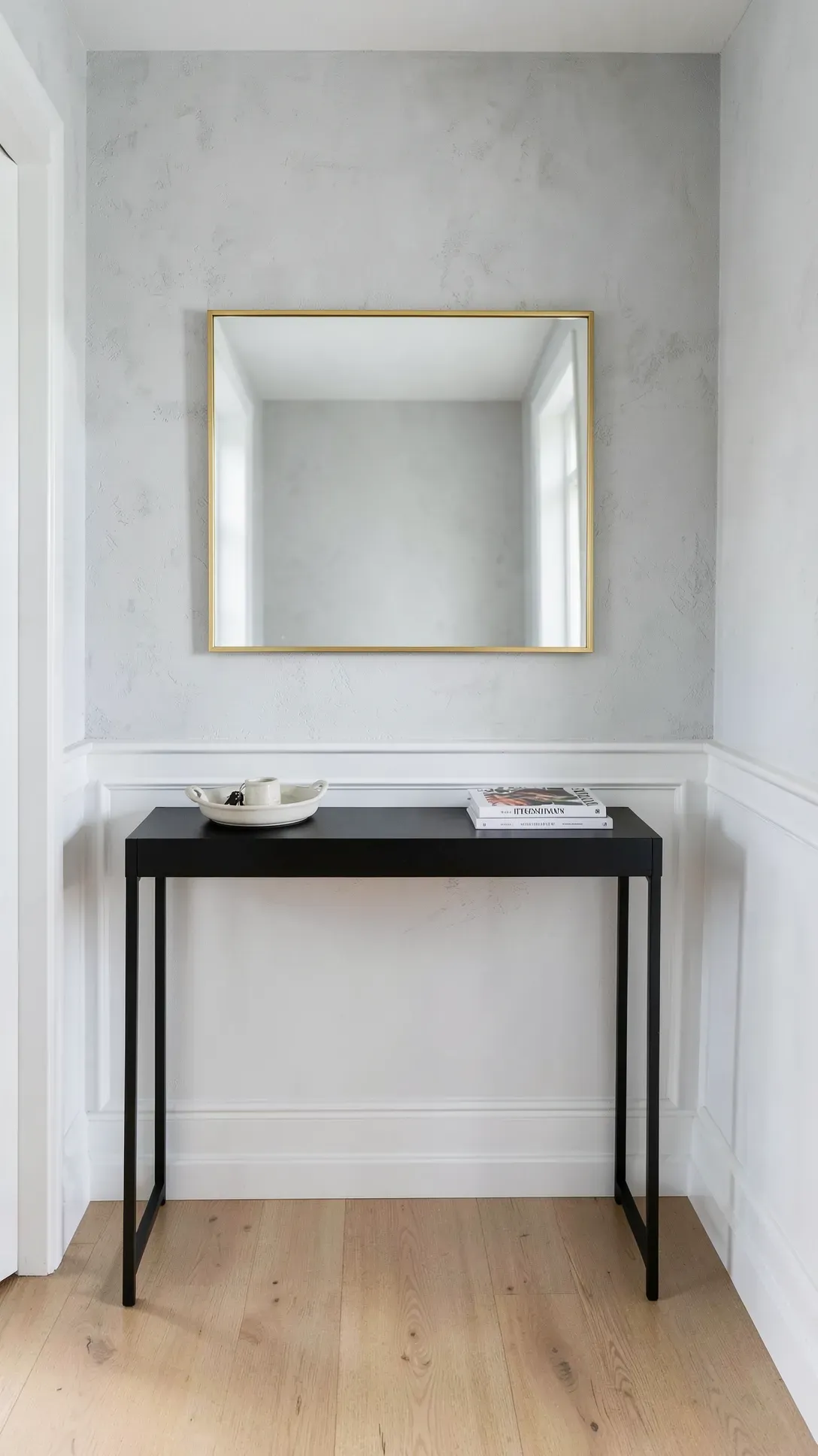 A horizontal gold-framed mirror hung over a narrow black console table in a bright, modern entryway.