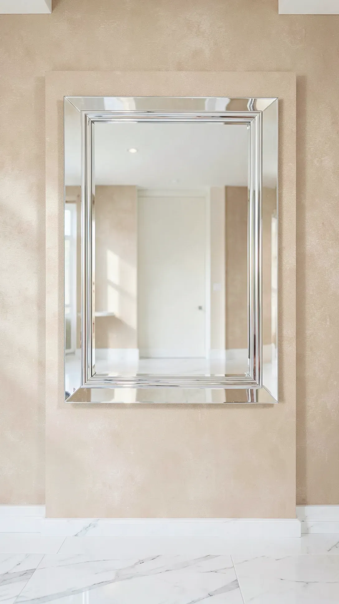 A frameless rectangular mirror with beveled edges on a champagne-colored textured wall.