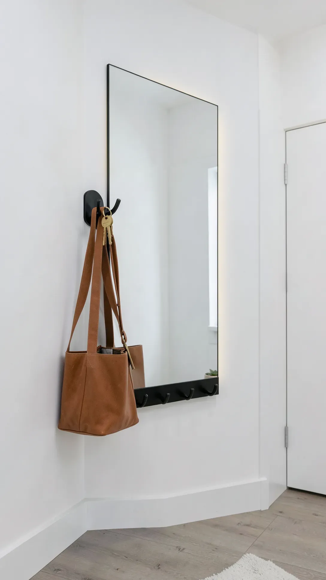 A functional rectangular mirror with attached black hooks holding a bag and keys in a white hallway.