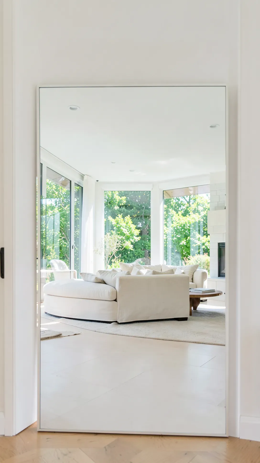 A wide horizontal mirror in an entryway reflects a bright, sunlit living room with a sofa.