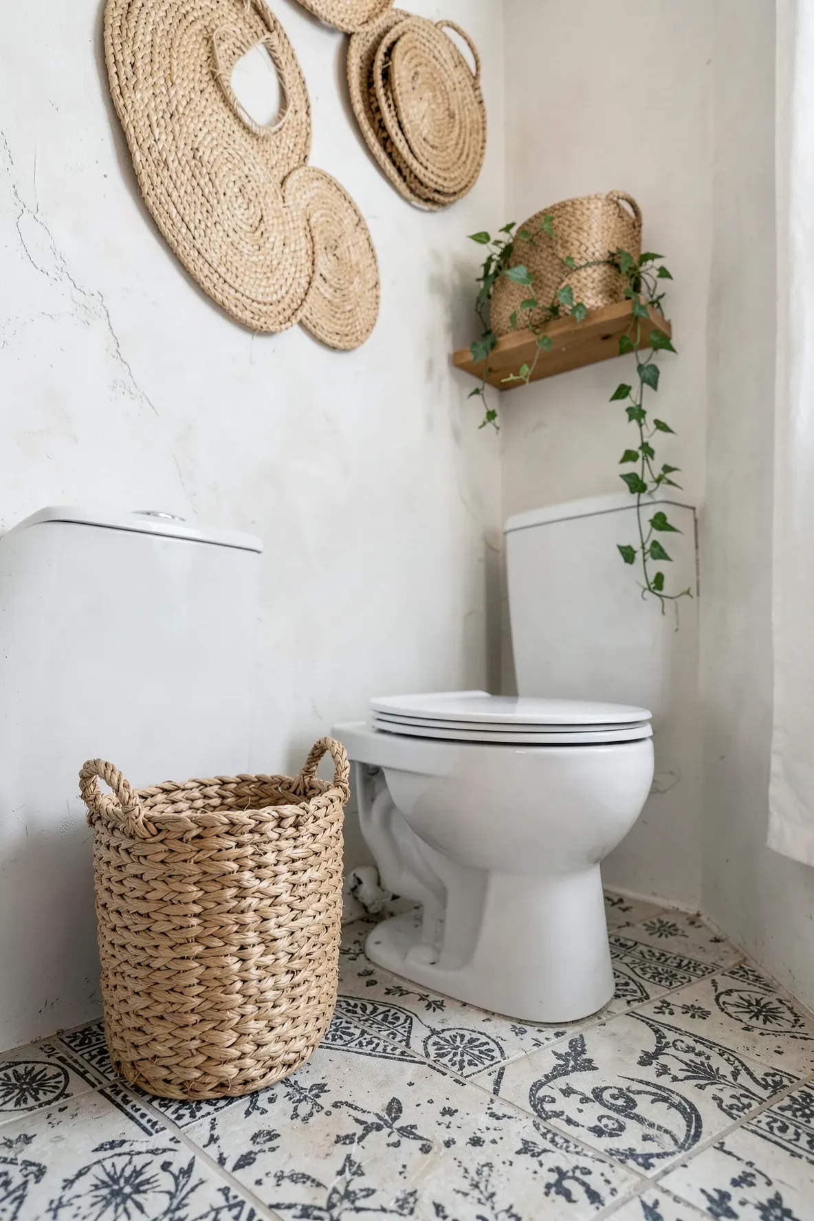 A woven seagrass wastebasket next to a white toilet in a bathroom with patterned tile floors.