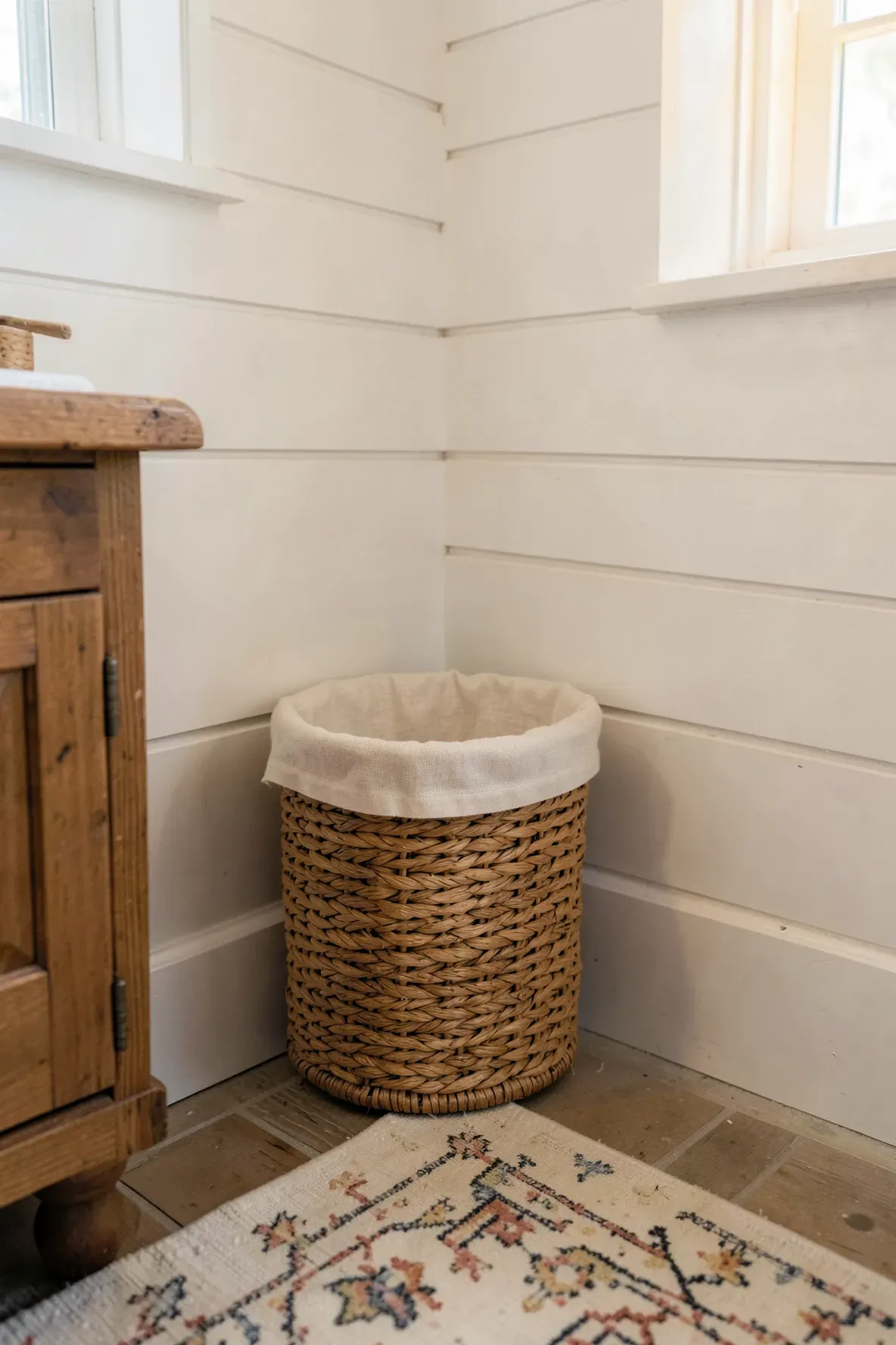 A woven rattan wastebasket with a fabric liner sitting on a patterned rug in a boho bathroom.