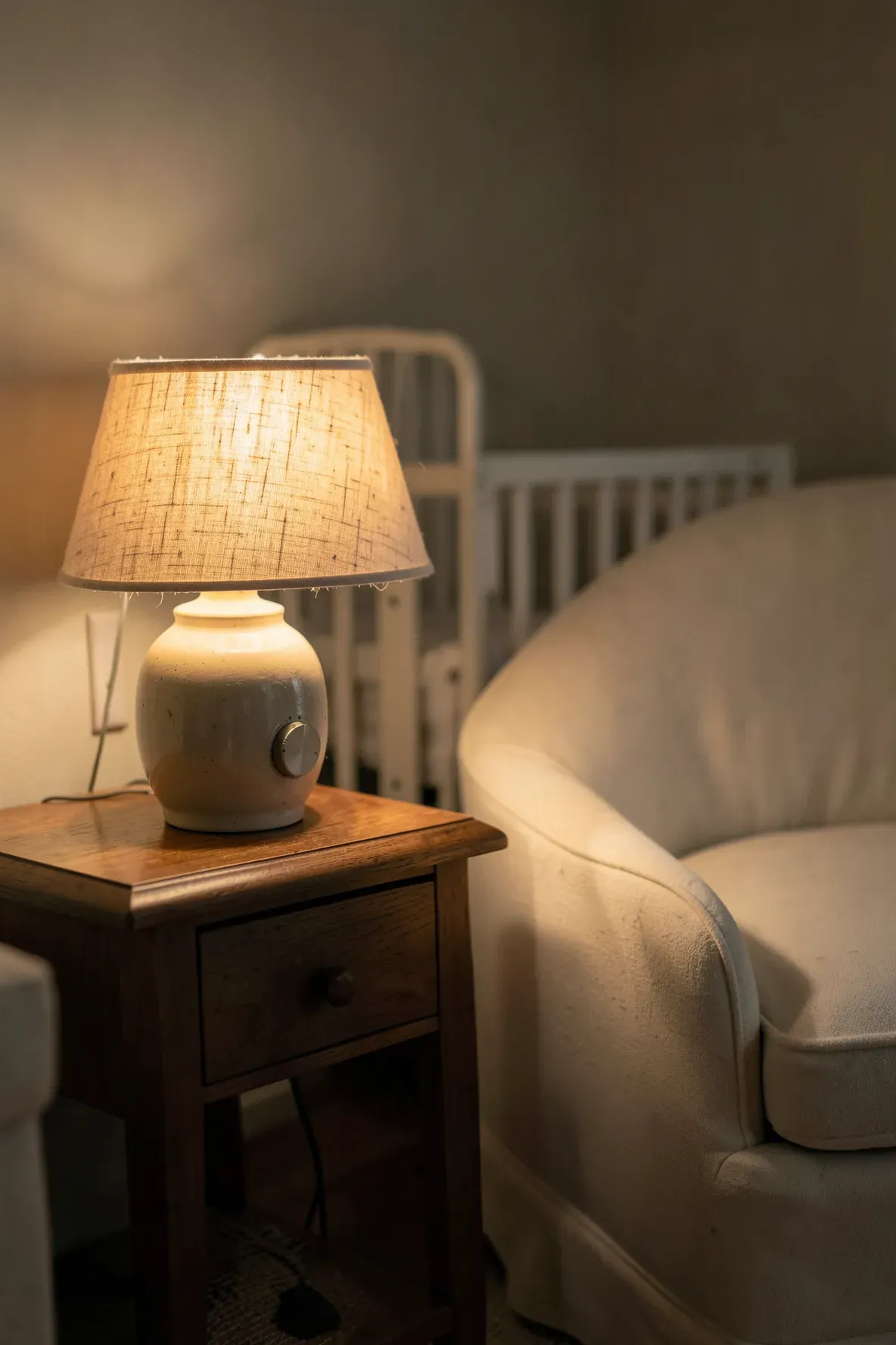 A warm, glowing table lamp on a side table next to a nursing chair in a dim nursery.