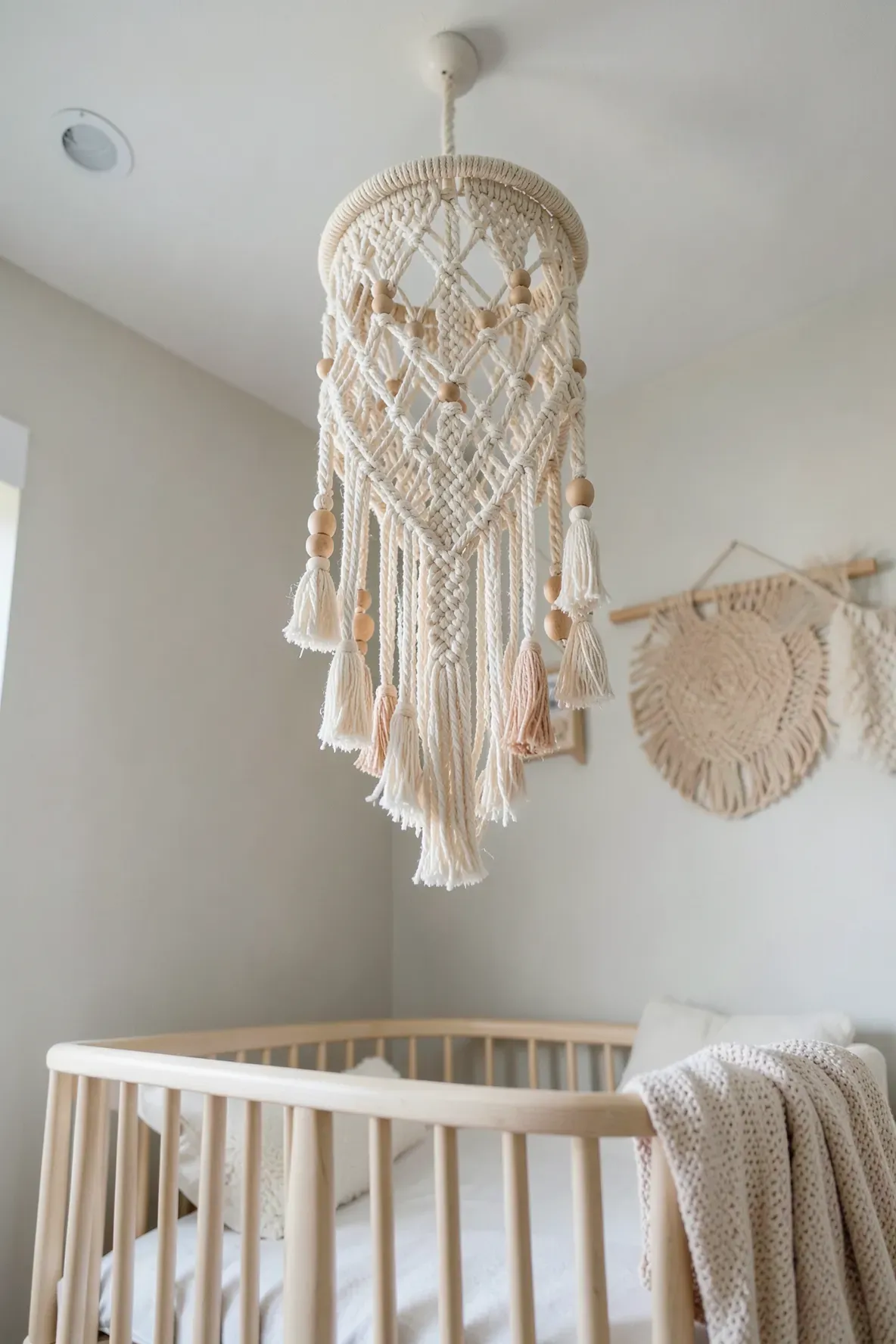 A cream macramé mobile hanging above a wooden crib in a boho-style nursery.