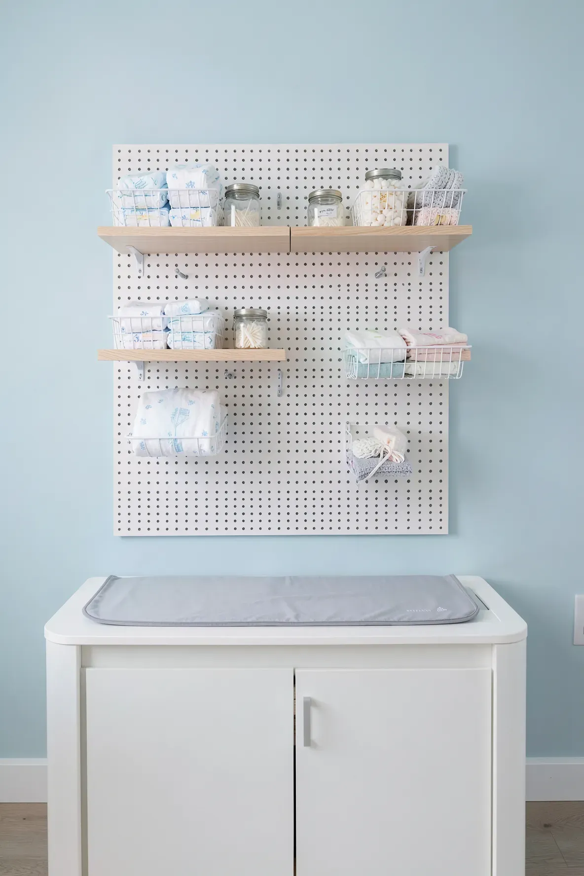 An organized white pegboard with shelves and baskets for diapering supplies above a changing table.