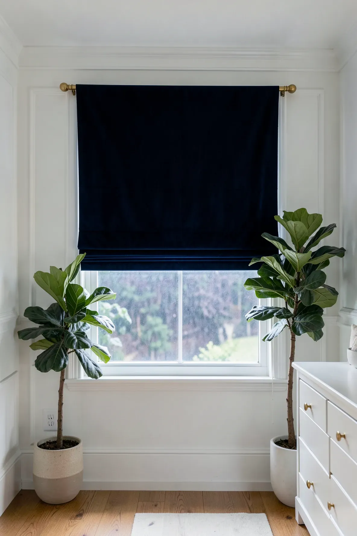 Navy blue velvet Roman shades installed on a window in a clean, white-walled nursery.