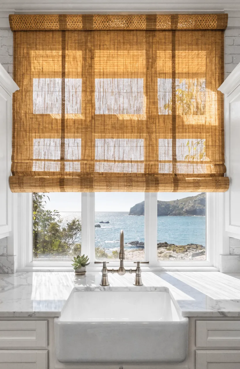 Honey-toned bamboo shades filtering sunlight over a white farmhouse sink in a coastal kitchen.