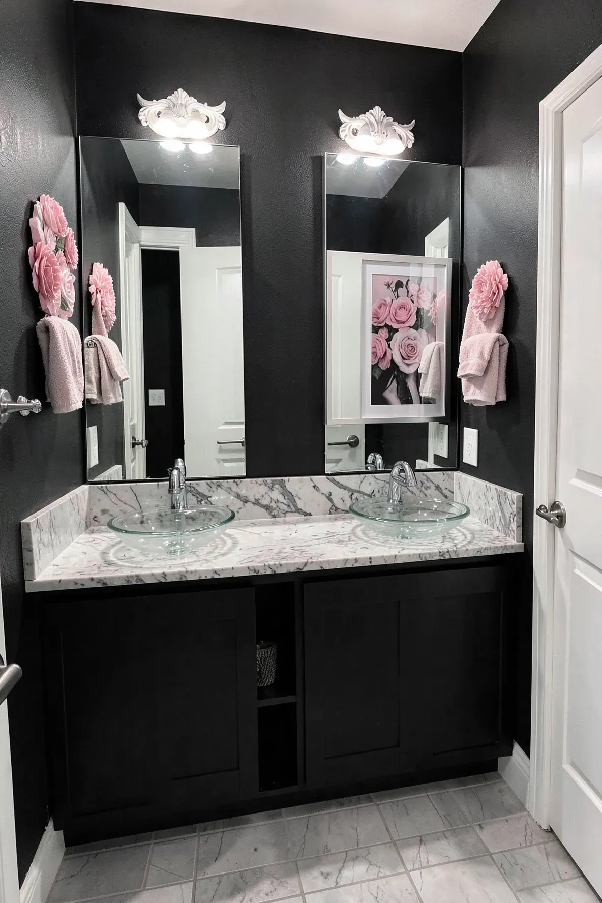 A modern bathroom vanity with a matte black backdrop, double glass vessel sinks, chrome faucets, dusty pink towels, and framed pink floral wall art with a white bull-head decor mount above the right piece.