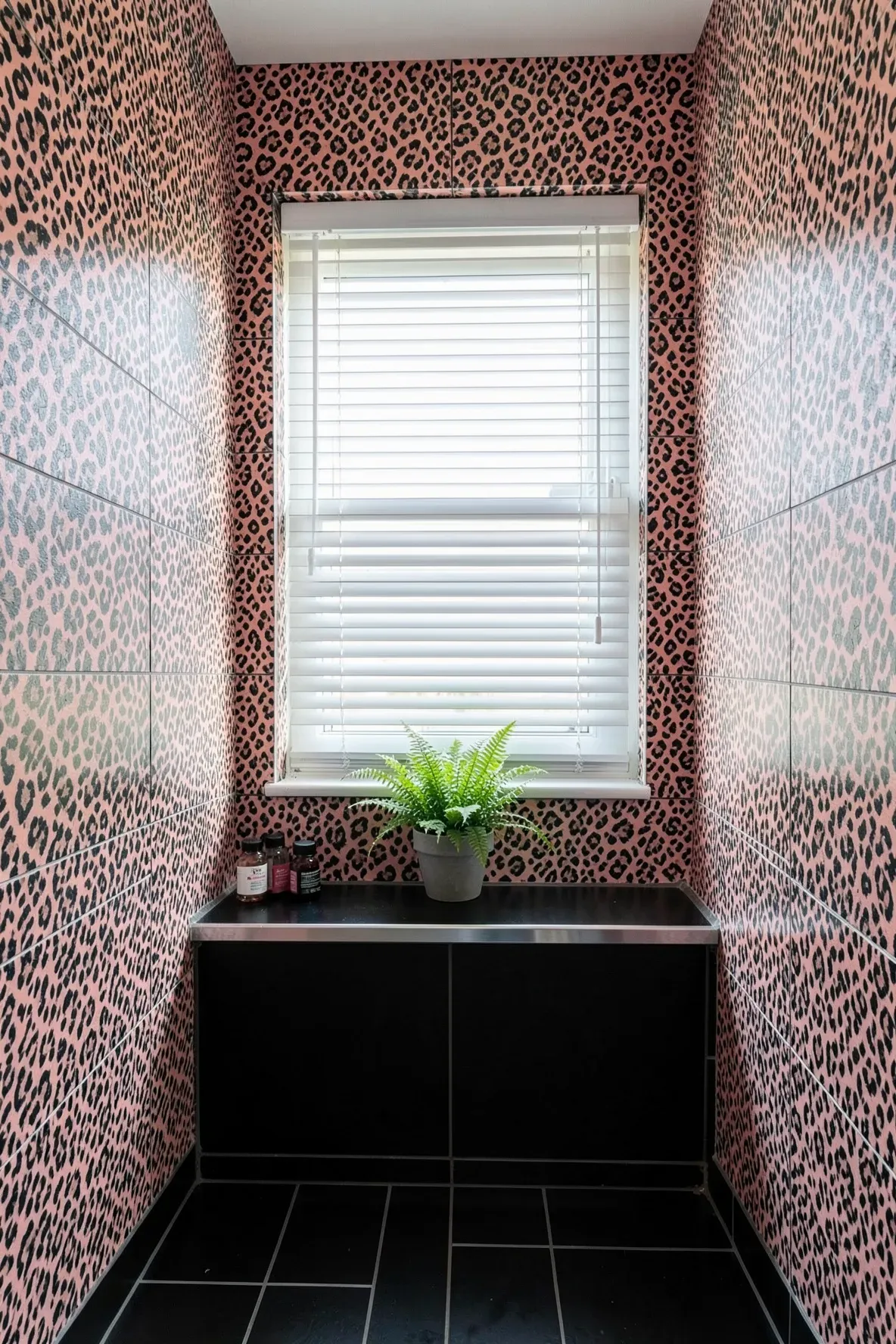 A modern corner bathroom with glossy black floor tiles, leopard-print wallpaper in dusty pink and coral tones around a window with white blinds, plus a small fern on the sill and minimal bottles.