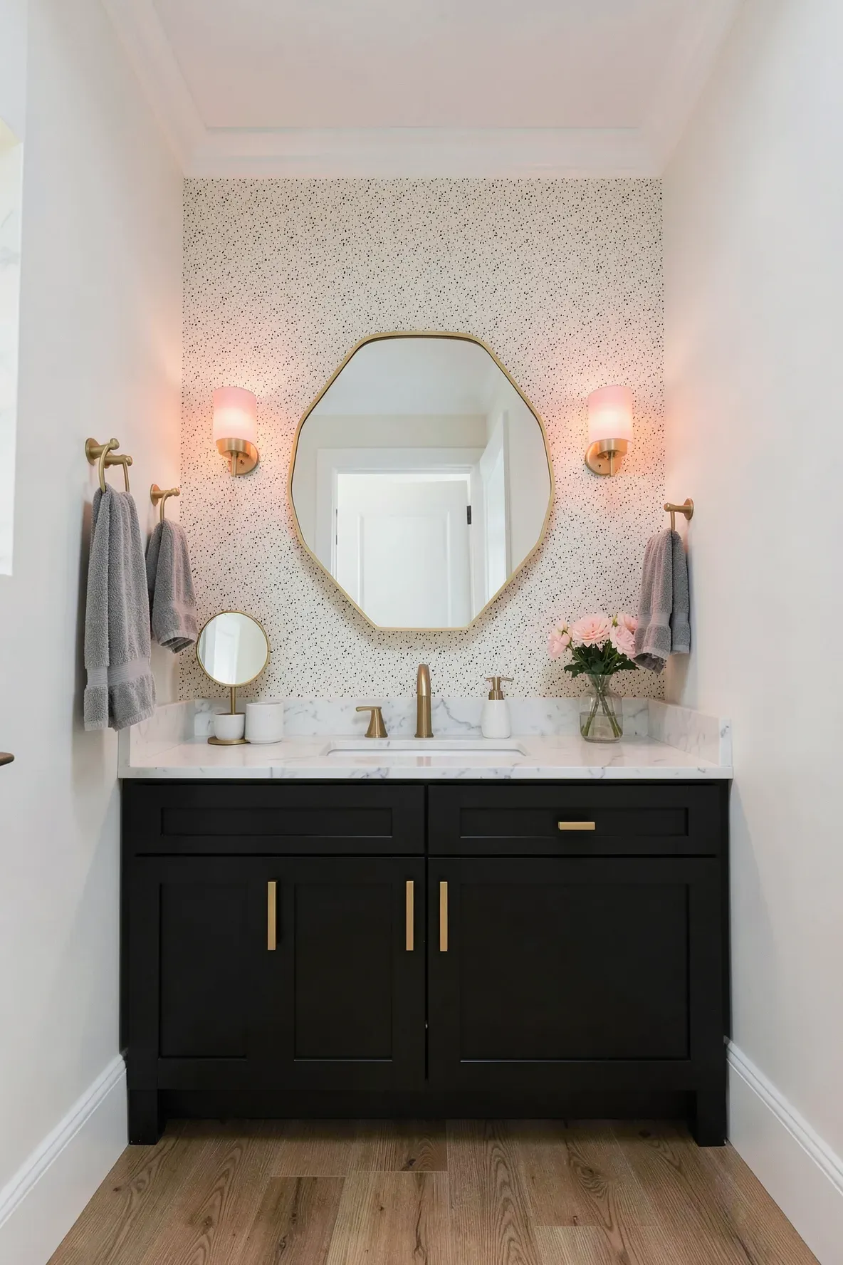 A modern vanity wall with matte black double cabinetry, a white marble-look countertop, a gold octagonal mirror, speckled black-and-cream wallpaper, gray towels on hooks, and warm pink ombré glass sconces glowing beside the mirror.
