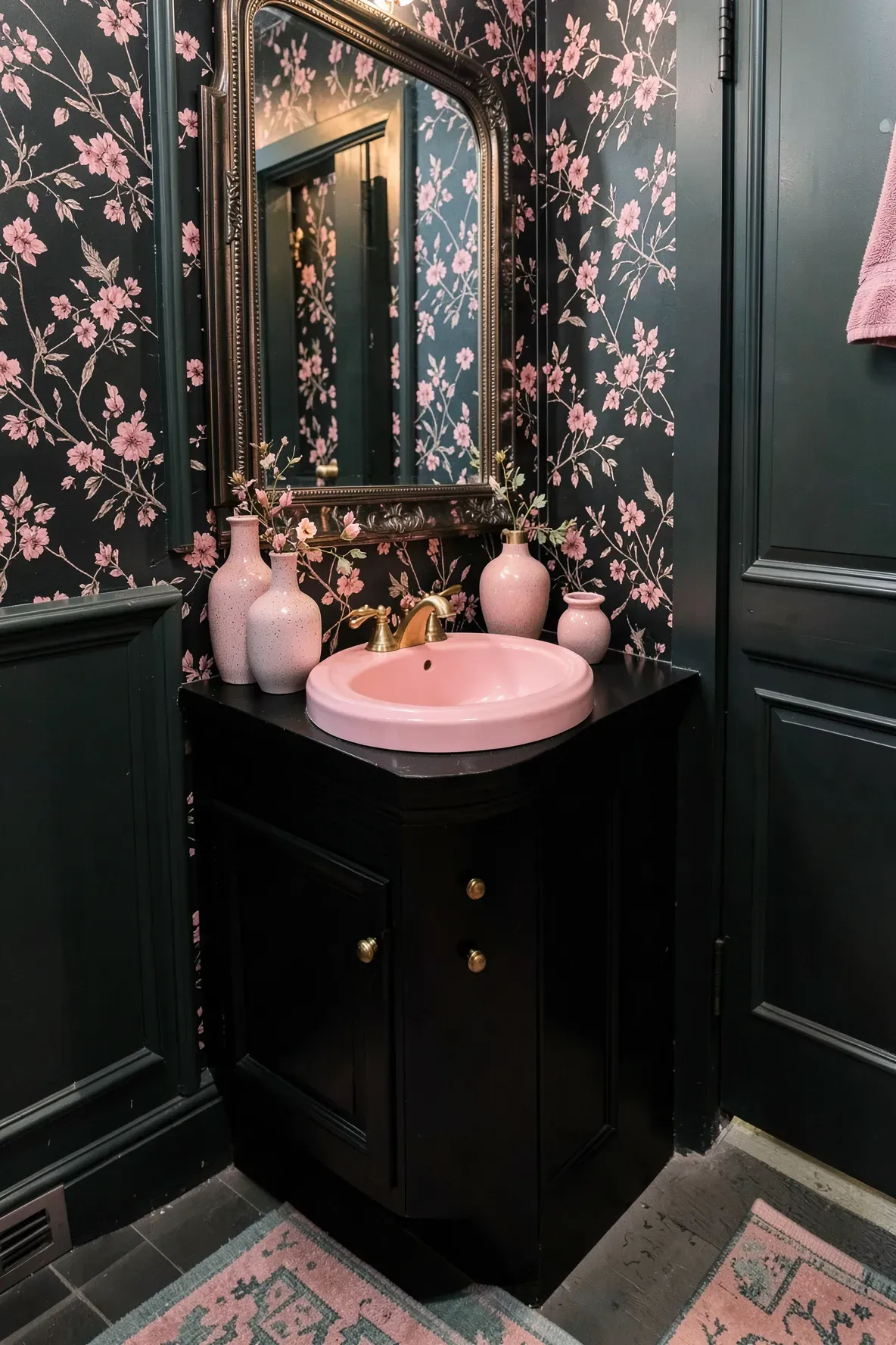 A moody corner bathroom with dense romantic floral wallpaper, a sleek black vanity, a rose-pink vessel sink, gold faucet hardware, a large ornate mirror, and a patterned rug under warm wall lighting.