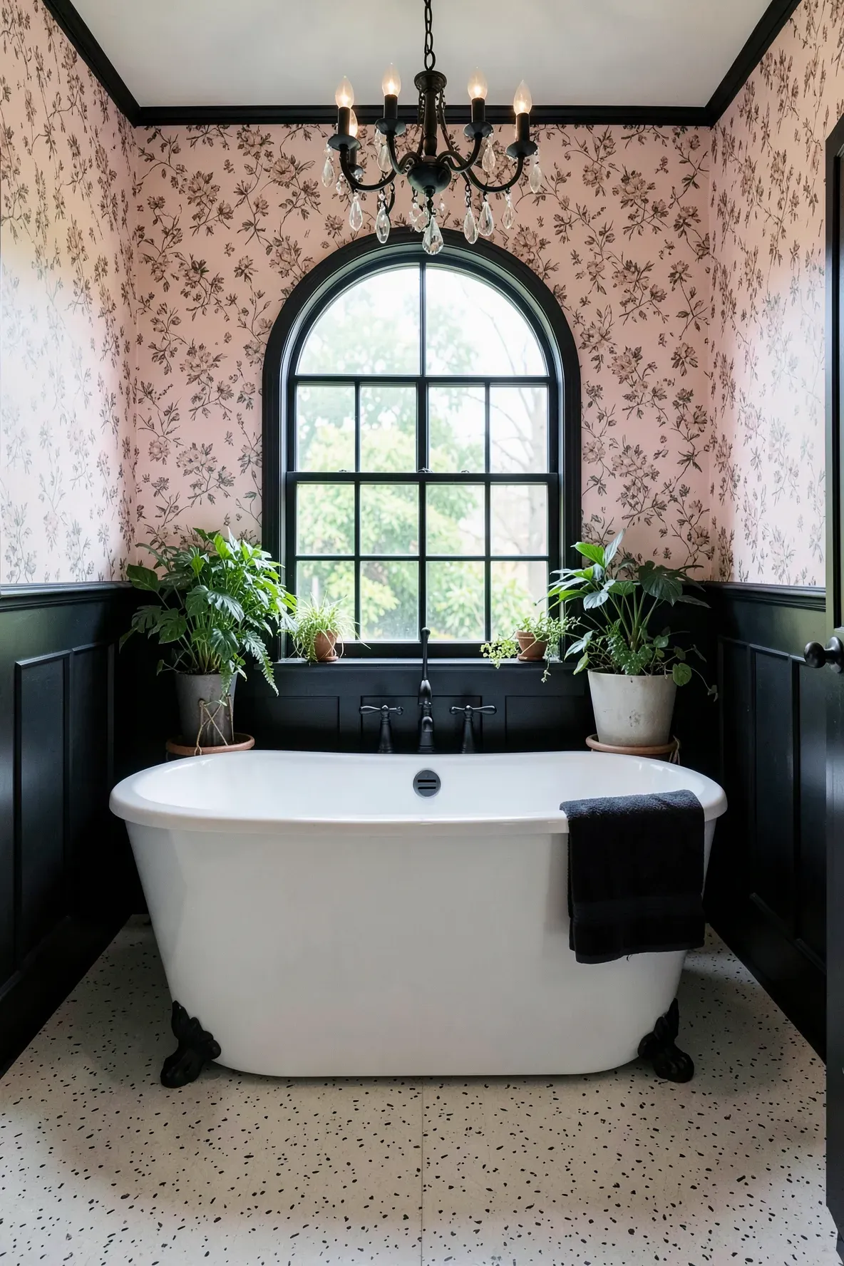 A symmetric modern-classic bathroom with blush floral wallpaper, a centered arched window with black trim, a white freestanding tub beneath, lush potted plants, and a black chandelier with crystal accents overhead.