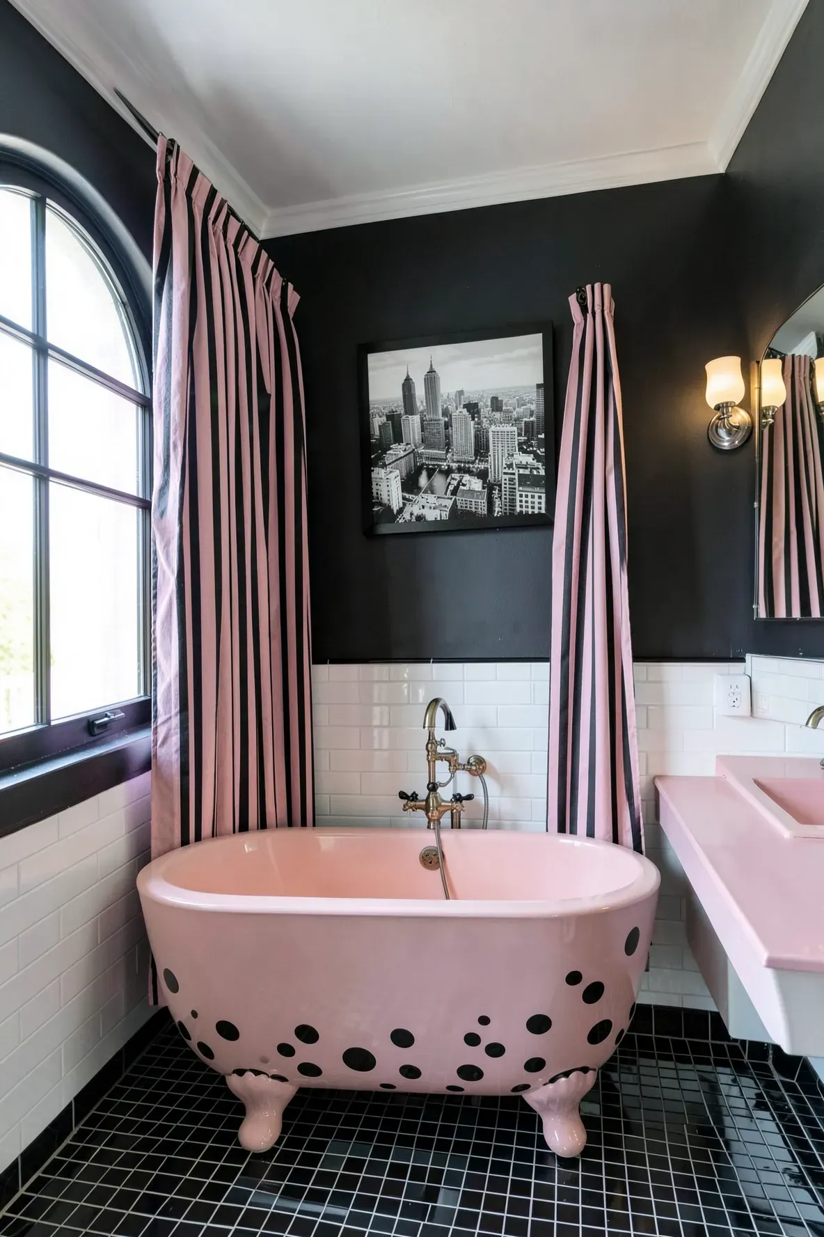 A modern glam bathroom with a blush-pink freestanding bathtub front featuring large black polka dots, black-and-blush striped curtains, a circular framed city photo above the tub area, and black-and-white accents.