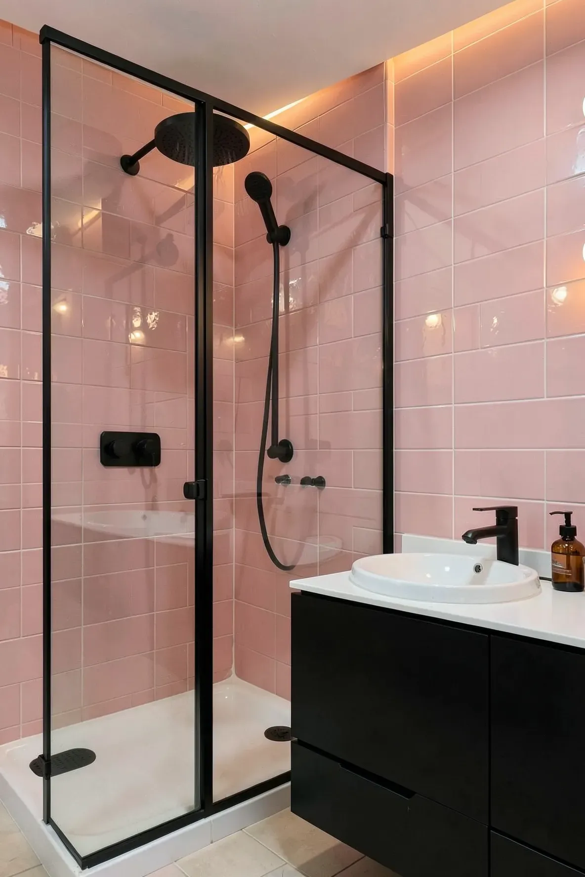 A modern bathroom with glossy rose-pink brick-style tiles, a corner walk-in shower with black-framed clear glass, a black-and-white vanity with a white vessel sink, and warm pendant lighting and a leafy plant accent.