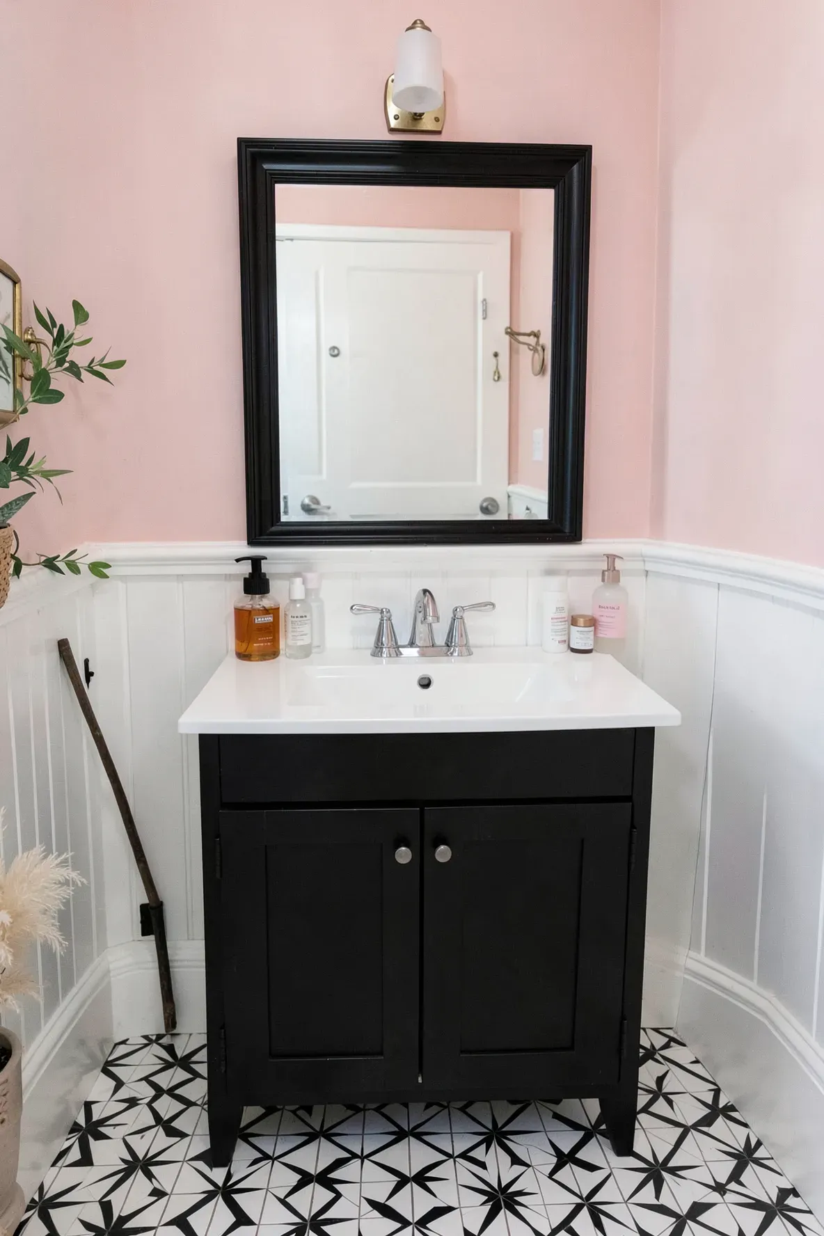 A small bathroom vanity scene with a matte black cabinet, a black-framed mirror, a chrome faucet over a crisp rectangular white undermount sink, and a black-and-white star patterned tile floor with a snake plant in the left corner.