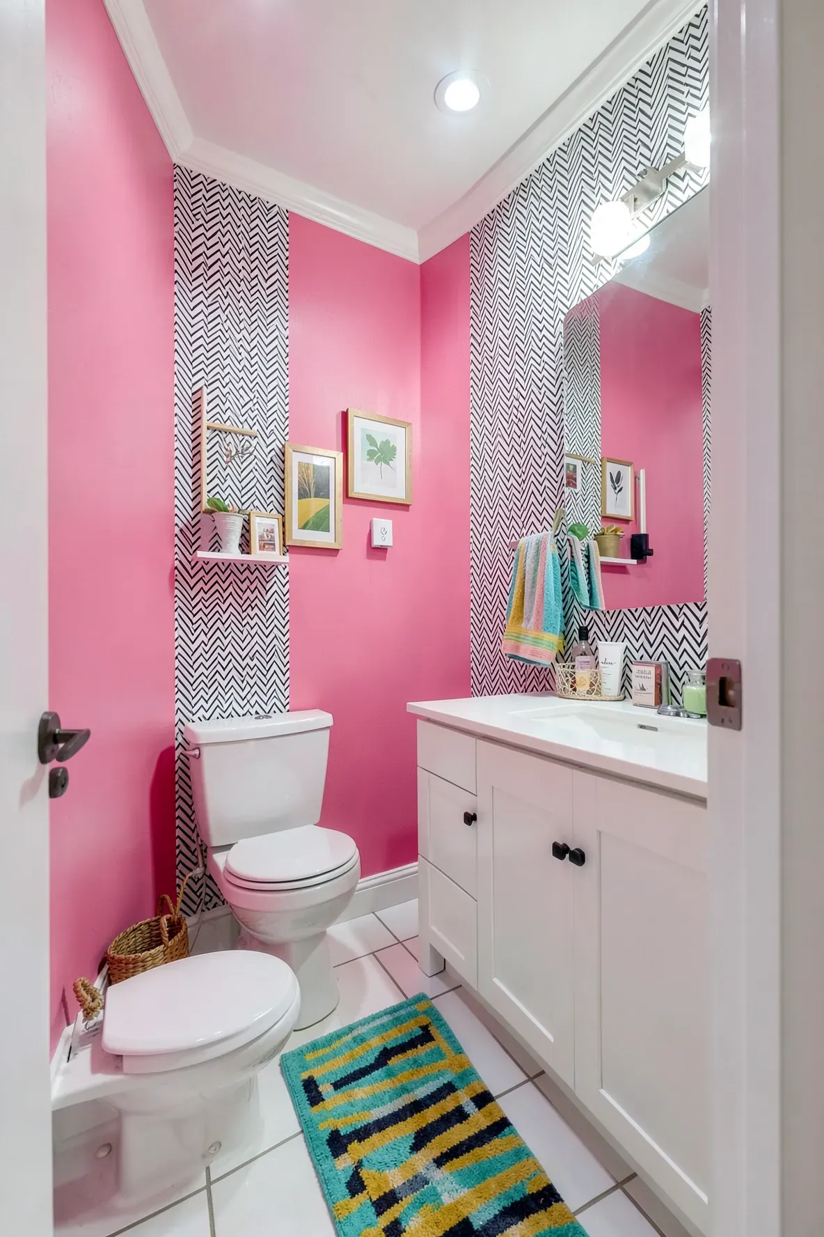 A small bathroom with hot pink painted walls, a black-and-white geometric wallpaper section behind the mirror, a white vanity, a modern white toilet, a geometric patterned rug with rainbow-like stripe accents, and colorful towels.