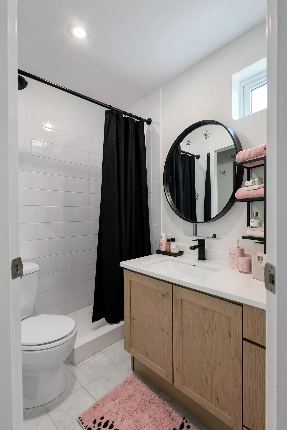 A bright modern bathroom with a white tiled shower, a black shower curtain, a light-wood vanity with matte black faucet hardware, a large mirror with rounded corners, blush-pink textiles, and a leopard-print rug in the foreground.