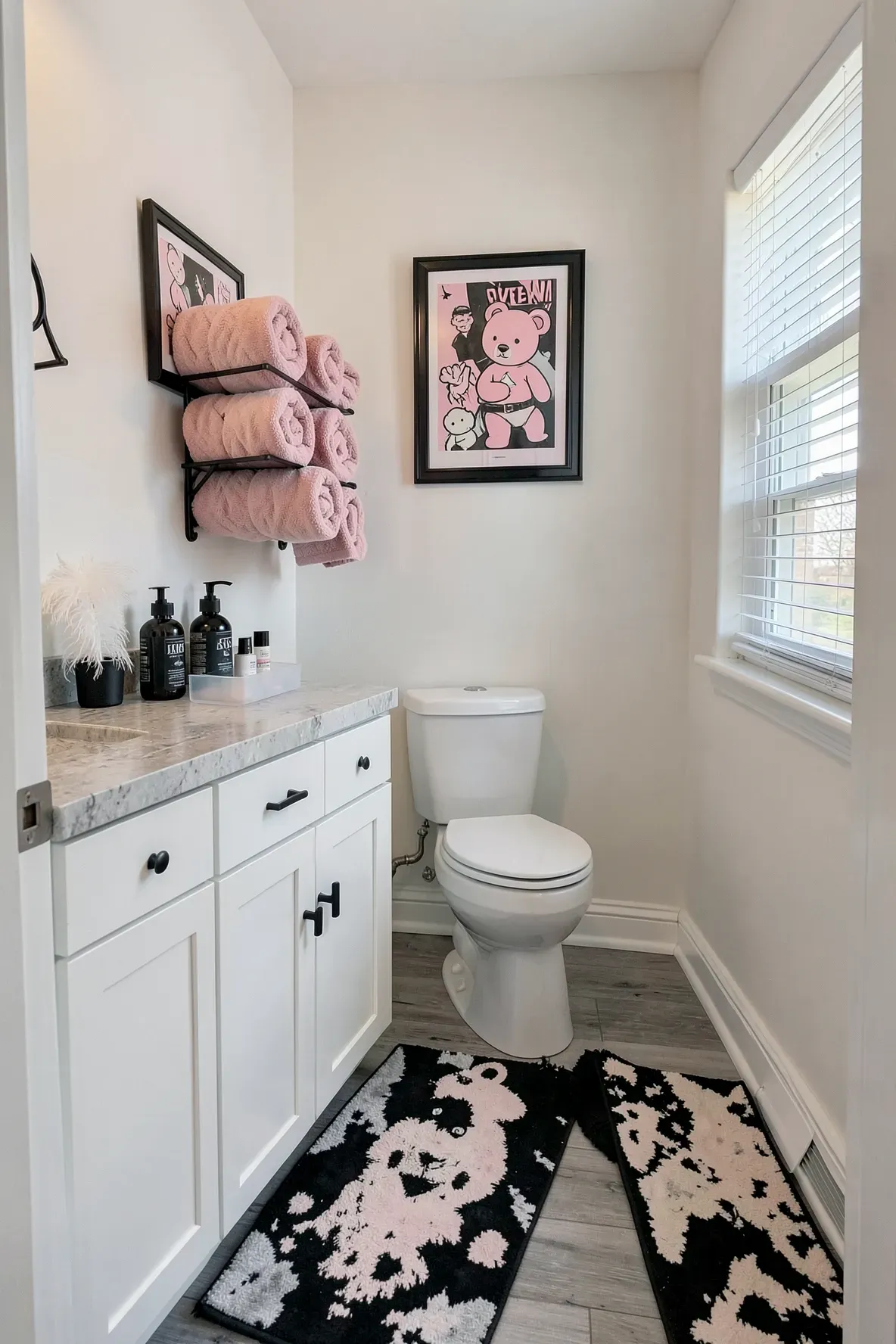 A modern bathroom with a white shaker-style vanity, black hardware, rolled blush-pink towels on a black tiered holder, two black-framed character wall posters, and daylight streaming from a window with blinds.