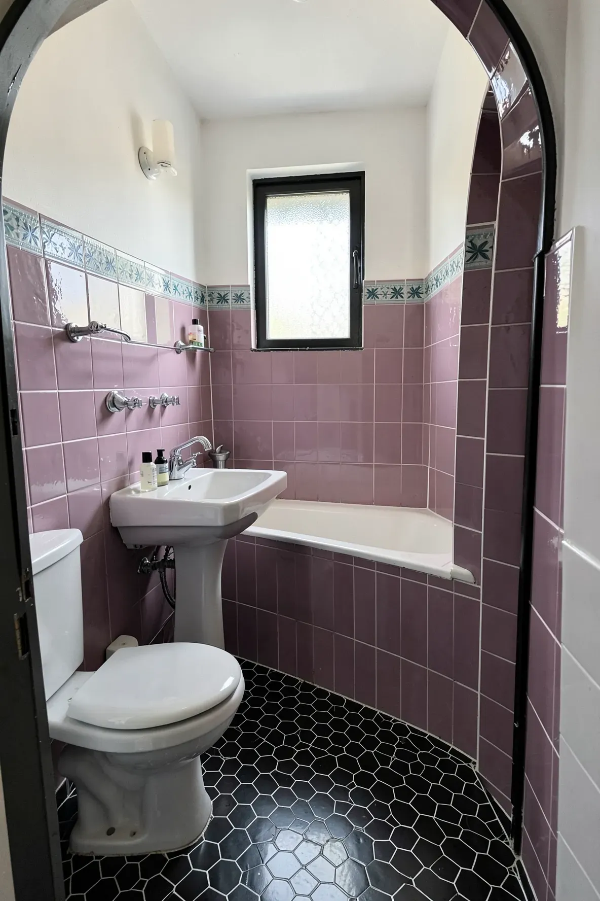 A compact tiled bathroom with matte black hexagon mosaic floor tiles, dusty mauve/pink tiled walls, a white pedestal toilet and sink, a tub-shower alcove with mauve surrounds, and a narrow teal border accent band.