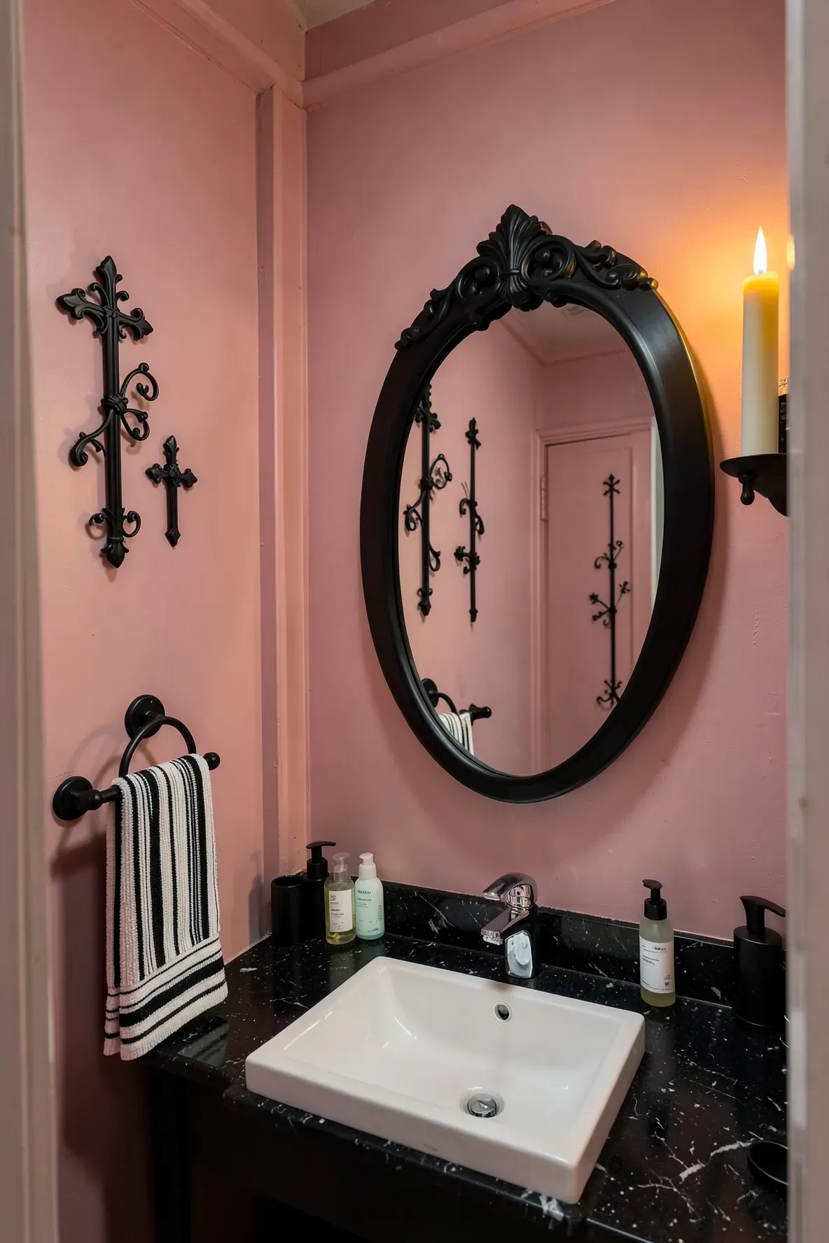 A cozy moody bathroom vanity area with a matte black ornate oval mirror, a dark countertop with a geometric white square sink, candlelit warmth, black baroque cross ornaments on the wall, and a striped black-and-white towel.