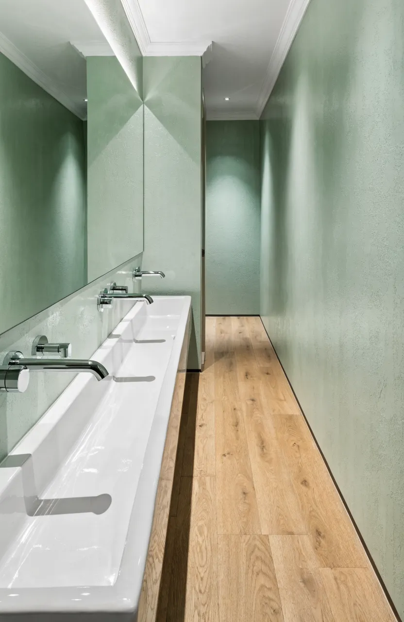 A slim white trough sink mounted on a green wall in a narrow bathroom with a clear walkway.