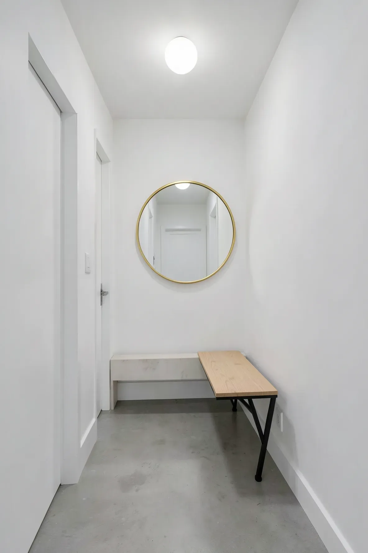 A circular gold mirror hanging above a narrow wooden bench in a bright white hallway.