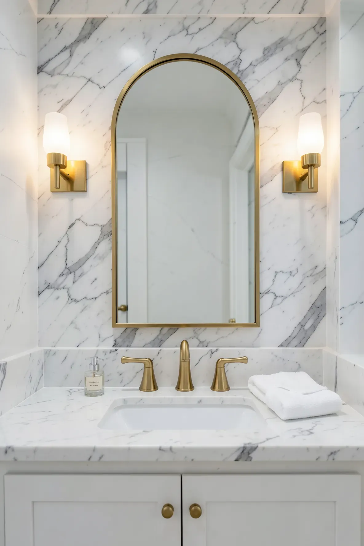 A gold arched mirror mounted above a white marble bathroom vanity with gold faucets and hardware.