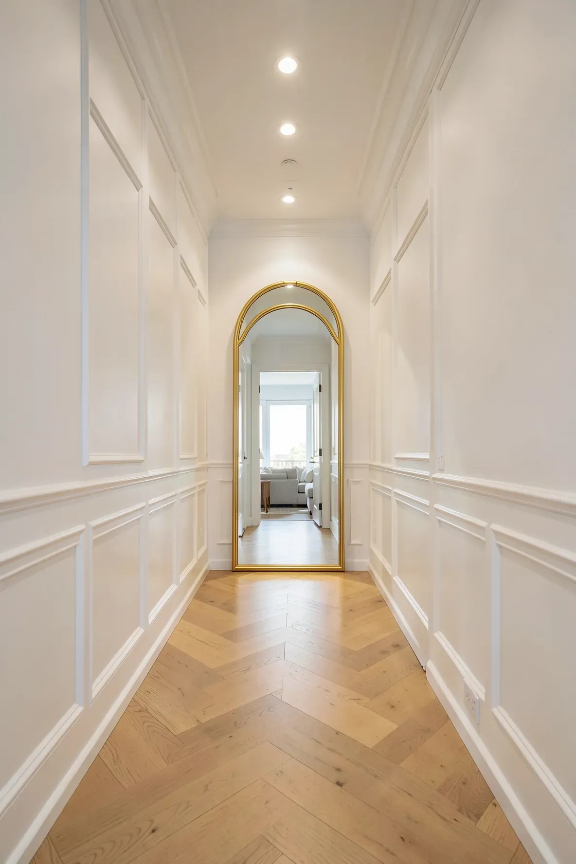 A long hallway with white walls and wood floors leading to a large gold arched mirror at the end.