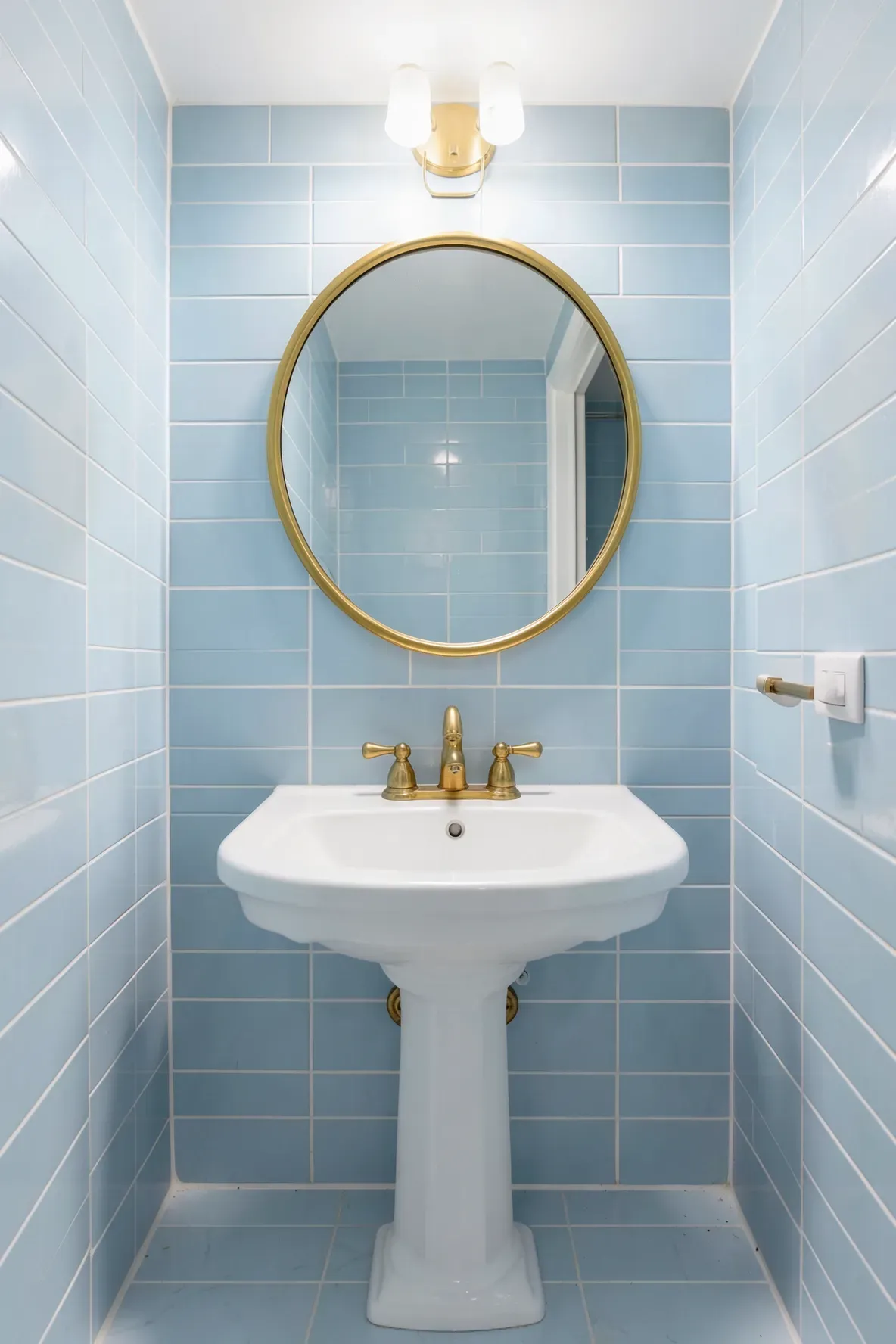 A circular gold mirror hanging above a white pedestal sink in a bathroom with light blue tiles.