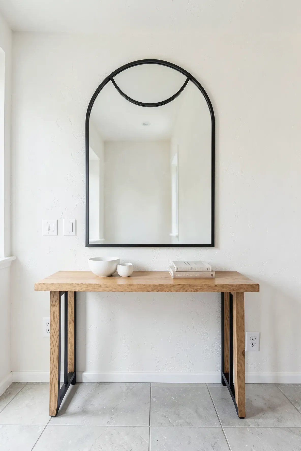 A black iron arched mirror hangs over a light wood console table with minimalist ceramic decor.