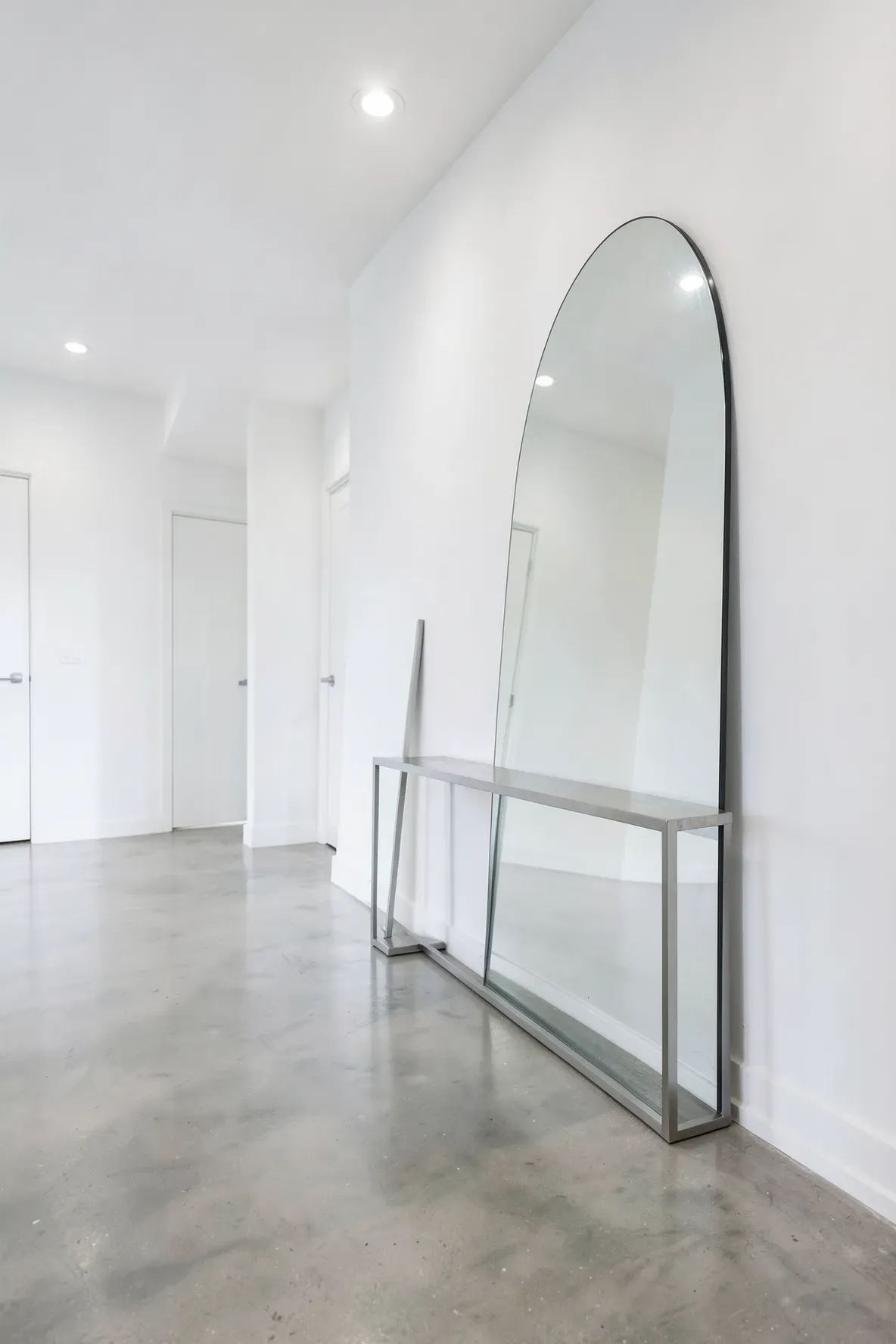 A large frameless arched mirror leans against a wall behind a narrow console table in a bright hall.
