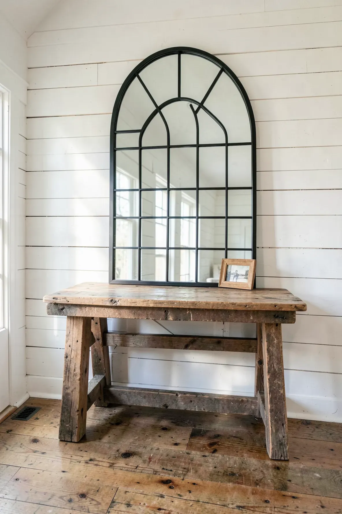 A black windowpane arched mirror sits above a rustic reclaimed wood table against shiplap walls.