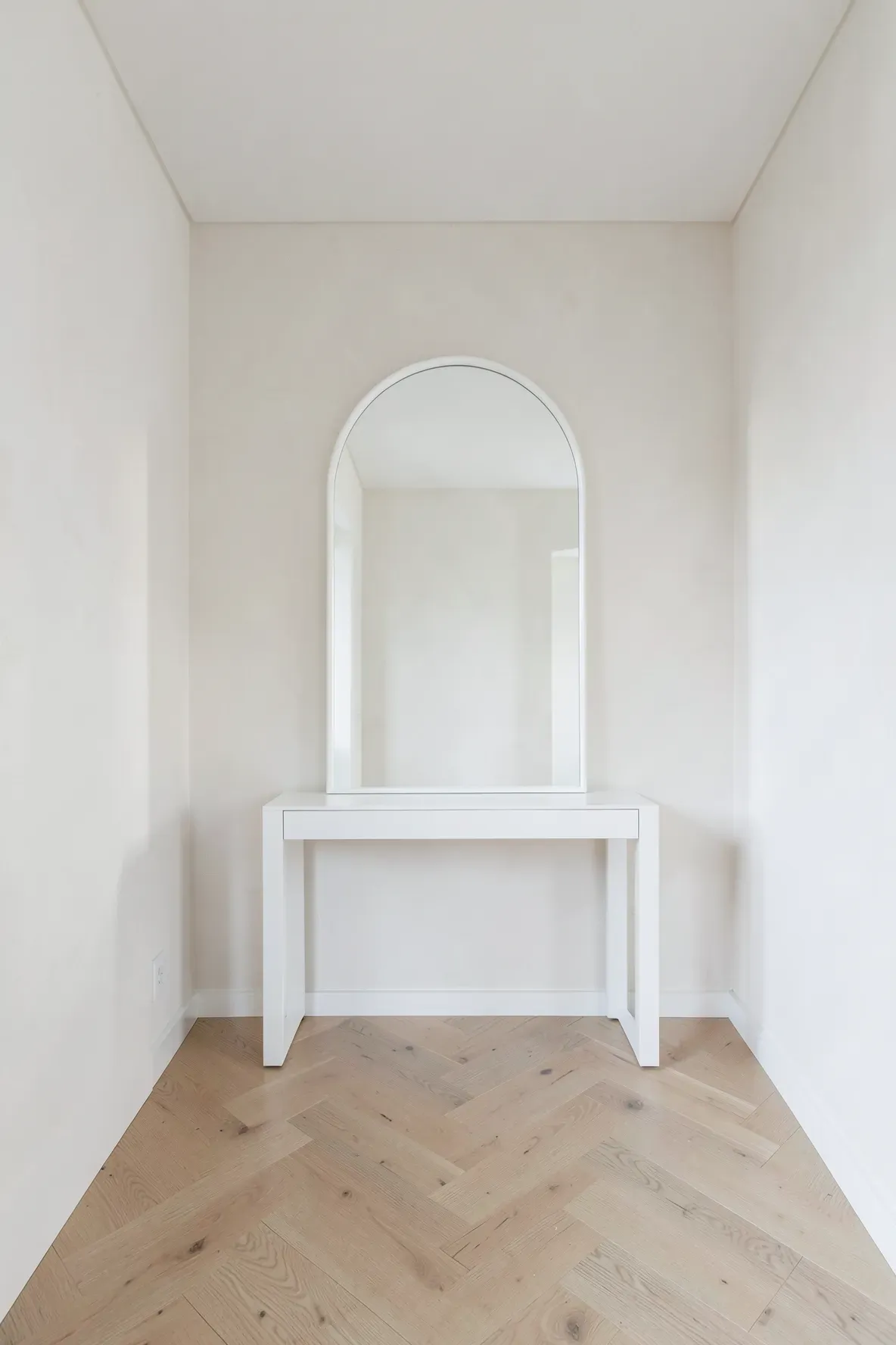 A matte white arched mirror mounted on a white wall above a matching slim console table.