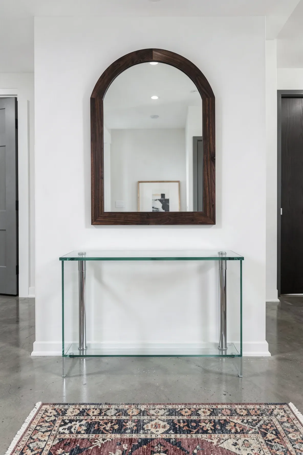 A dark wood arched mirror hanging above a clear glass entryway table in a modern hallway.