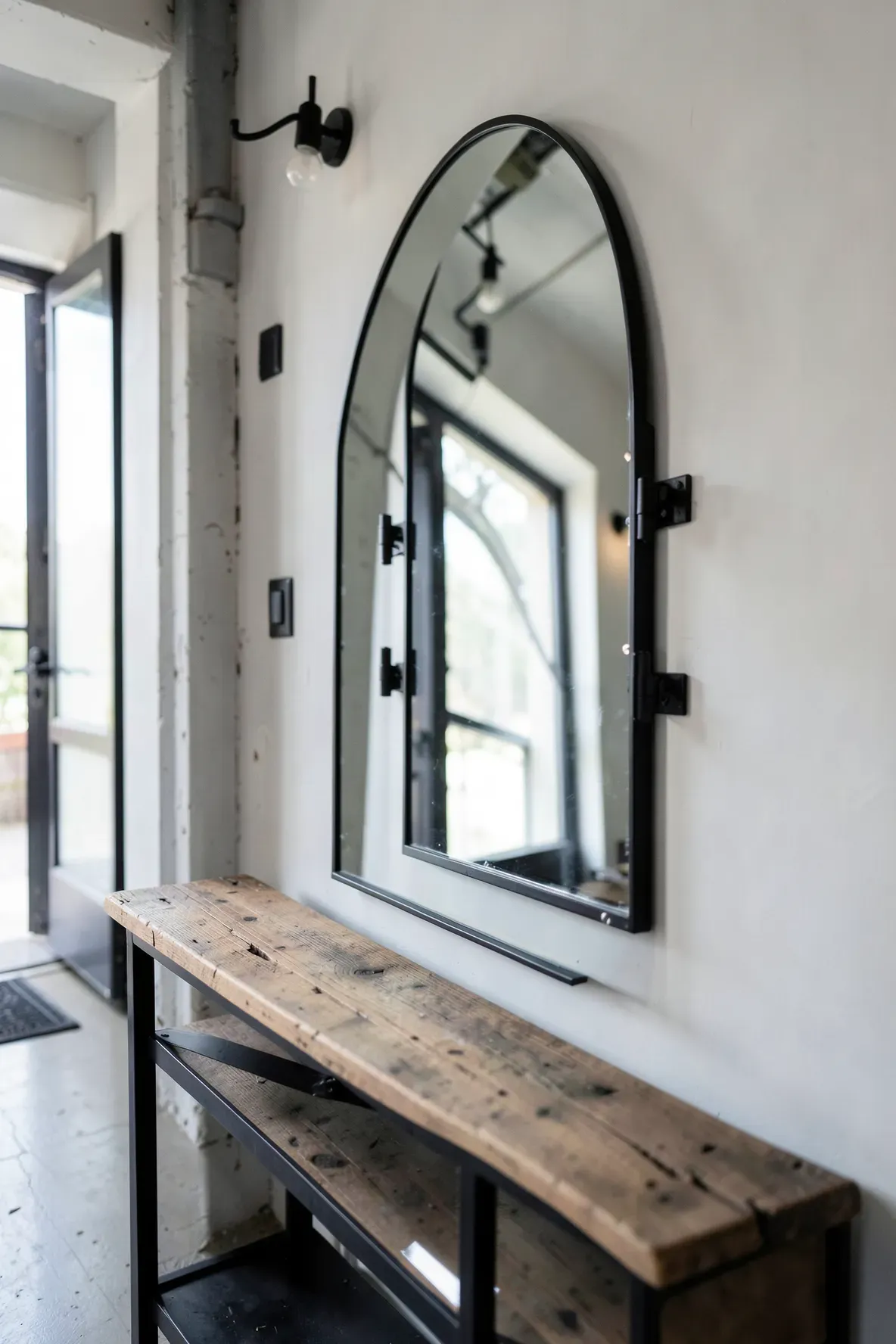 An industrial black metal arched mirror with pivot hinges mounted above a hallway table.
