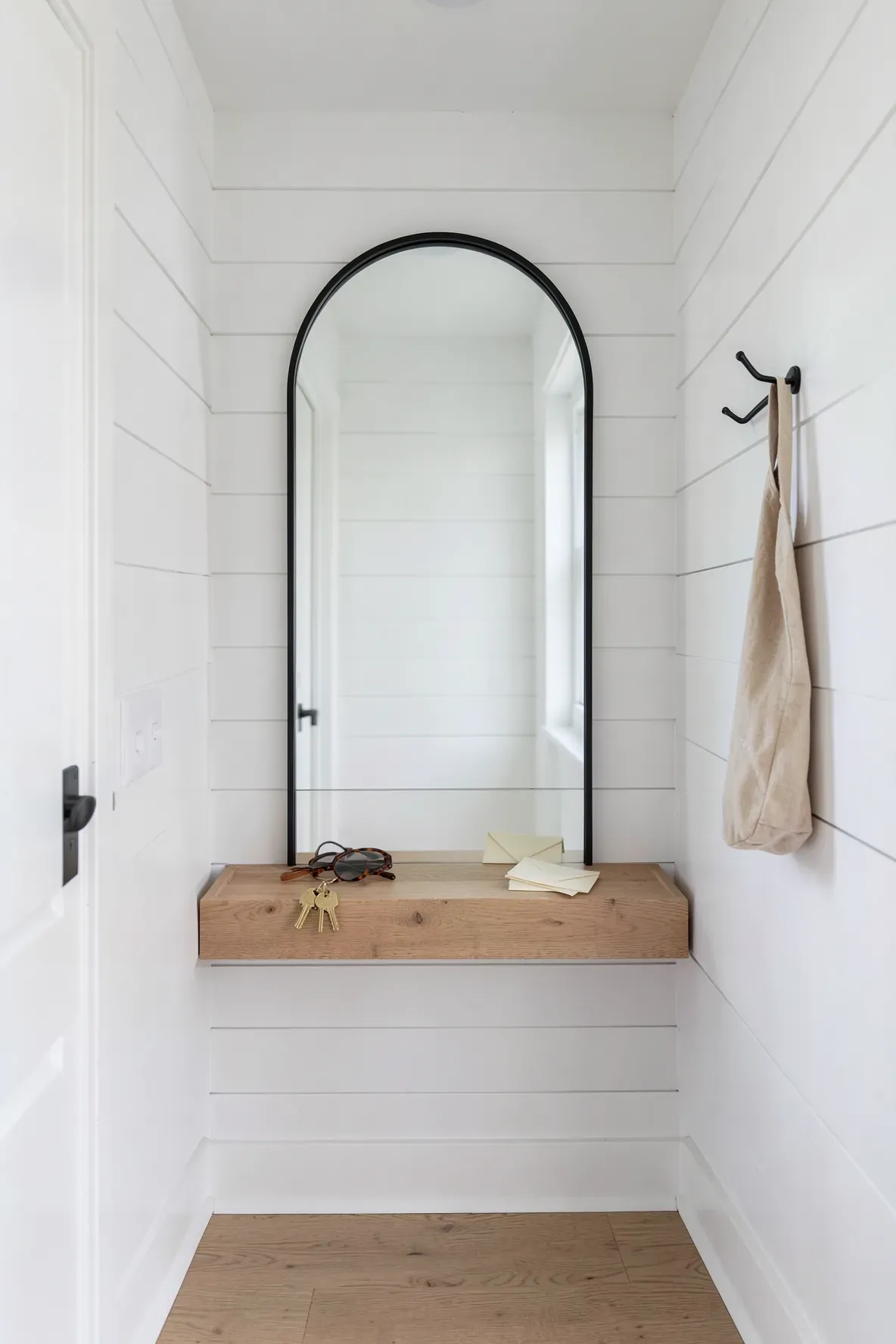 A black arched mirror with a built-in wooden shelf holding keys and sunglasses in a bright hallway.