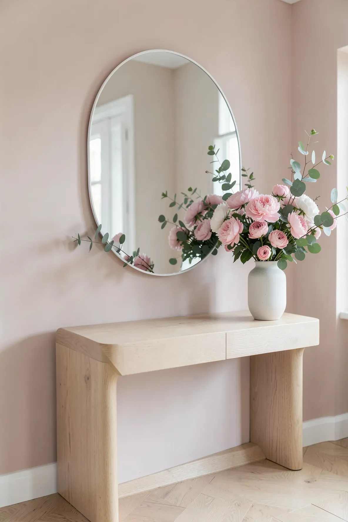 A white arched mirror on a light wood table with a tall pink floral arrangement on one side.