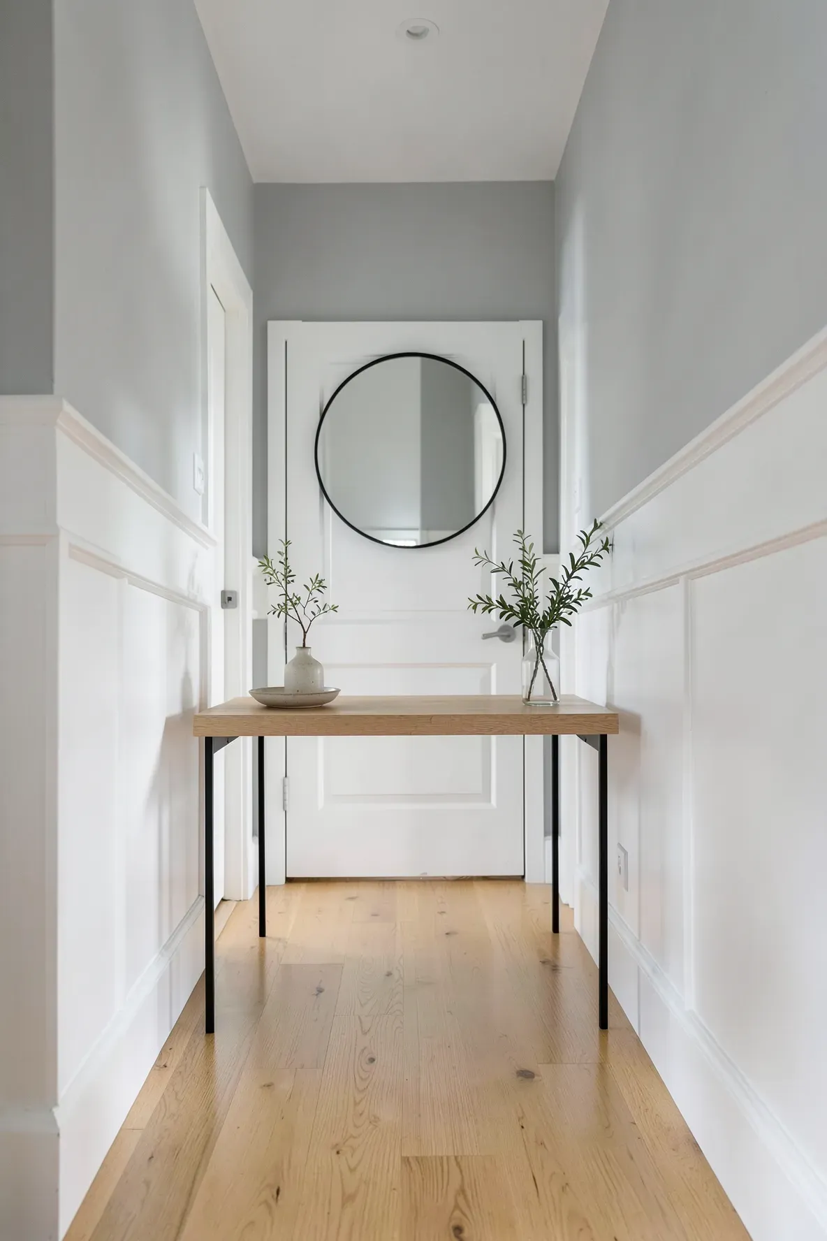 A slim wooden console table with a large round mirror in a bright, narrow white hallway.