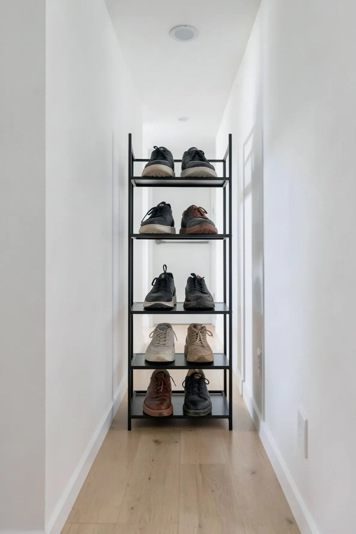 A slim matte black metal shoe rack against a white wall in a narrow modern hallway with wood floors.