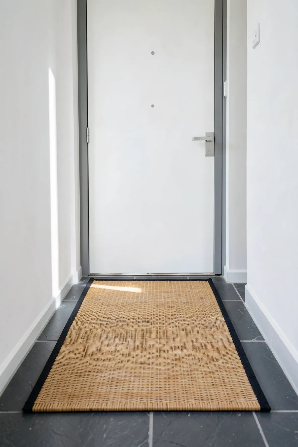 A thin, natural bamboo mat with a black border on a gray tile floor in a narrow entryway.
