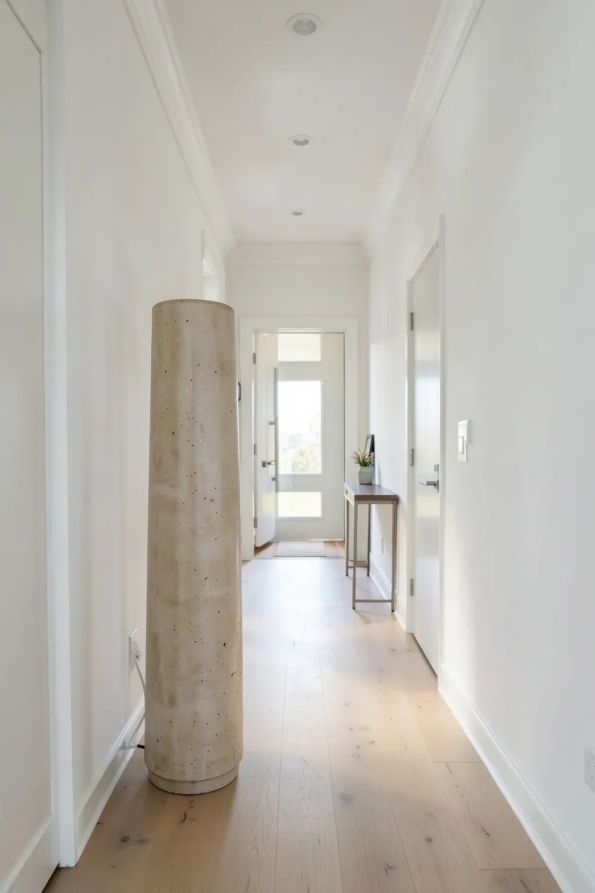 A tall cream ceramic umbrella stand in a bright, narrow modern hallway with wood floors.