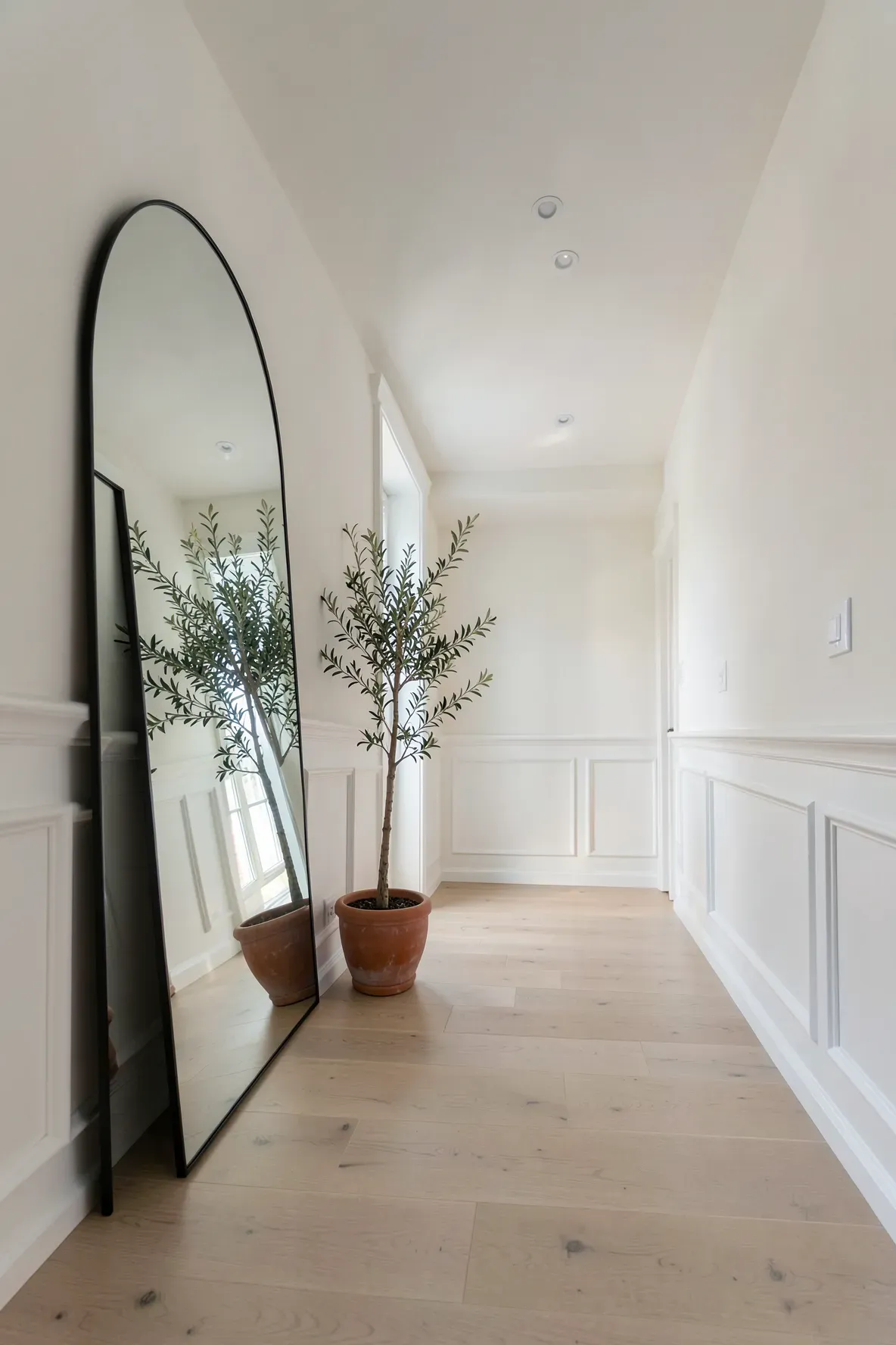 A large arched floor mirror leaning against a white wall in a bright, modern hallway with oak floors.