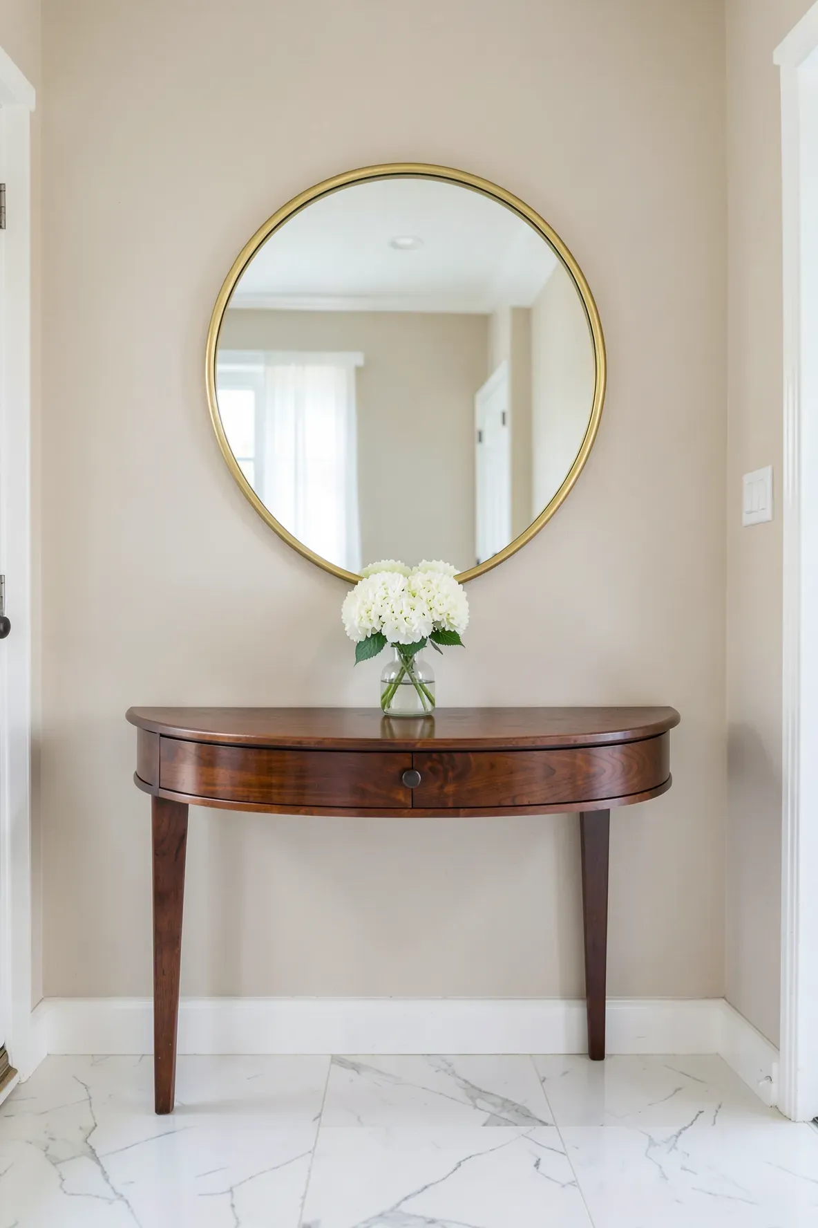 A round brass mirror hanging above a mahogany demi-lune table in an entryway.