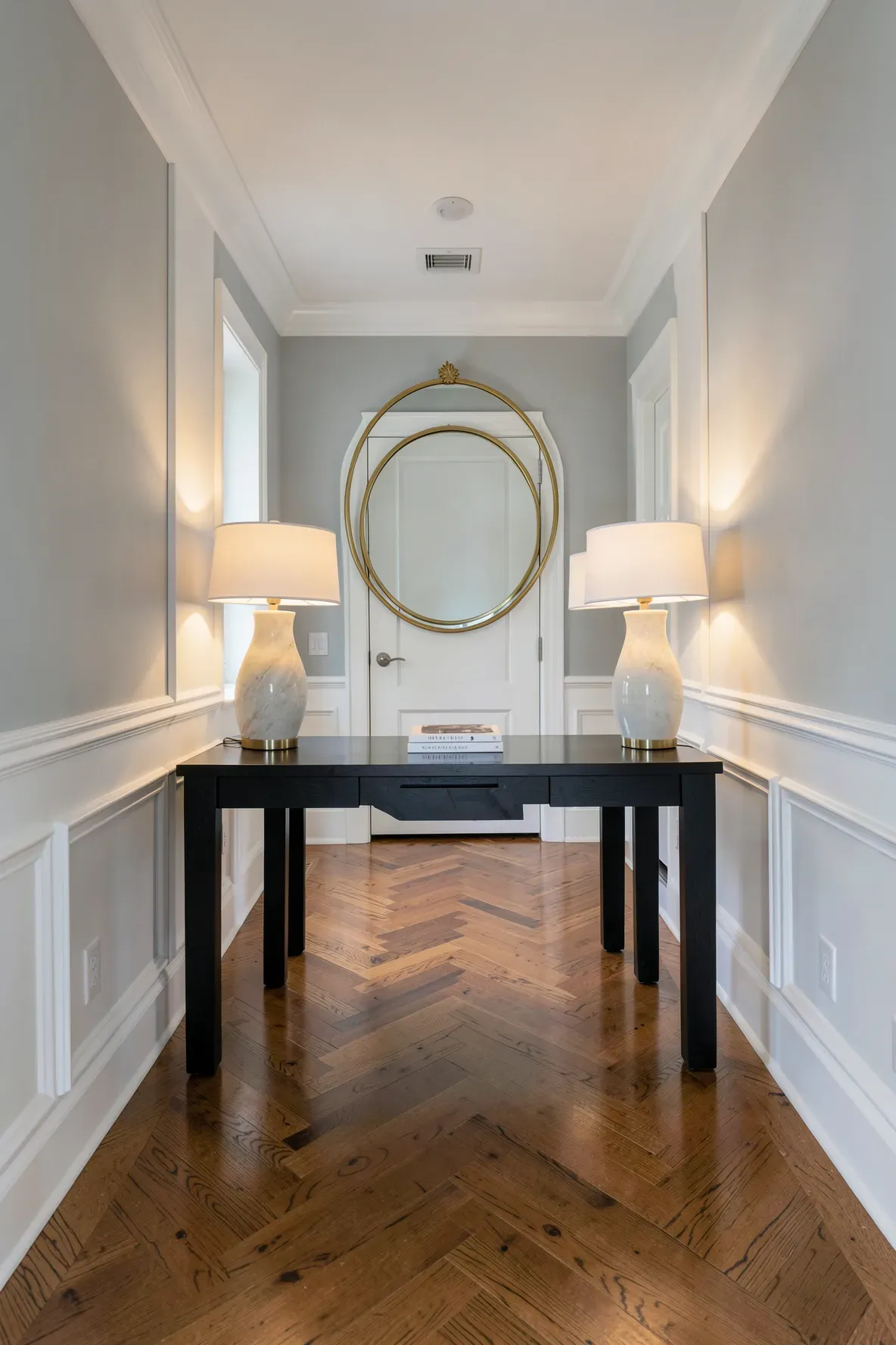 A symmetrical hallway featuring a black console table with two matching lamps and a gold mirror.