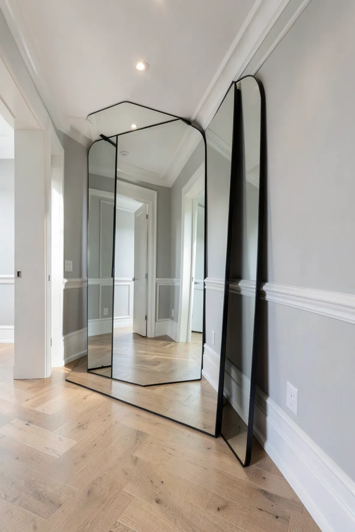 A large black-framed leaner mirror reflecting a bright, modern hallway with herringbone wood floors.