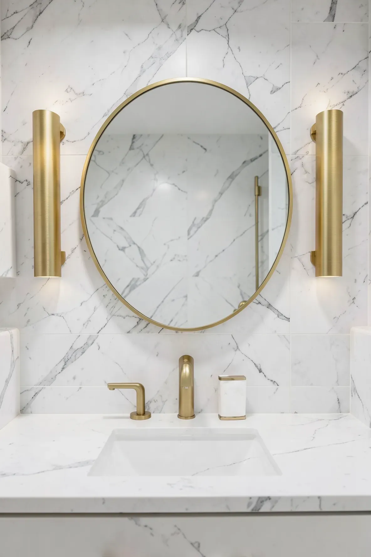 Modern brushed gold wall sconces flanking a circular mirror in a white marble bathroom.