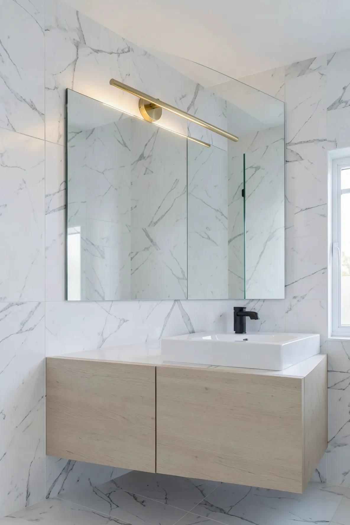 A minimalist slim brass bar light installed above a mirror in a modern white marble bathroom.