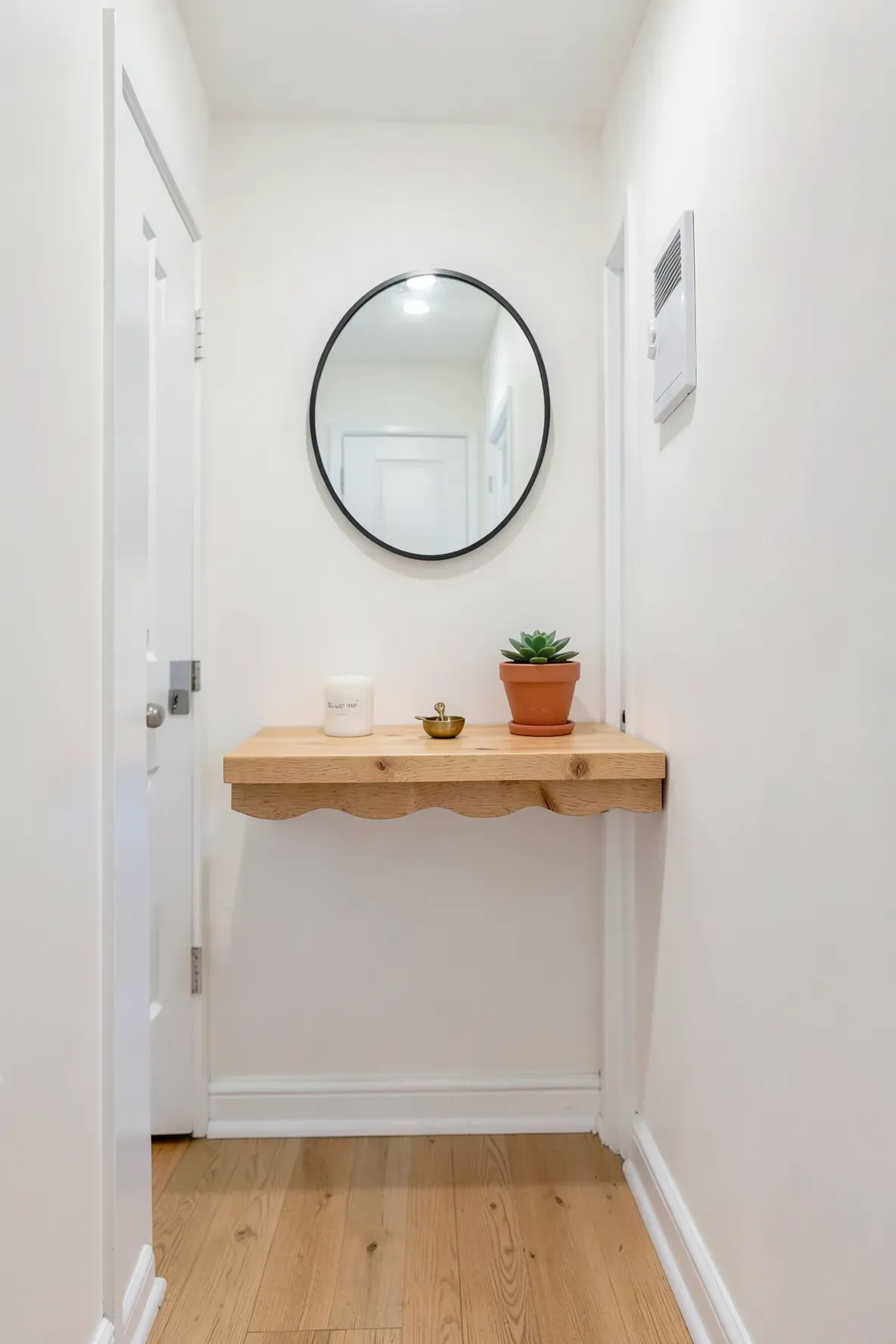 A small pine scalloped shelf in a narrow white hallway holding a key bowl and a small plant.