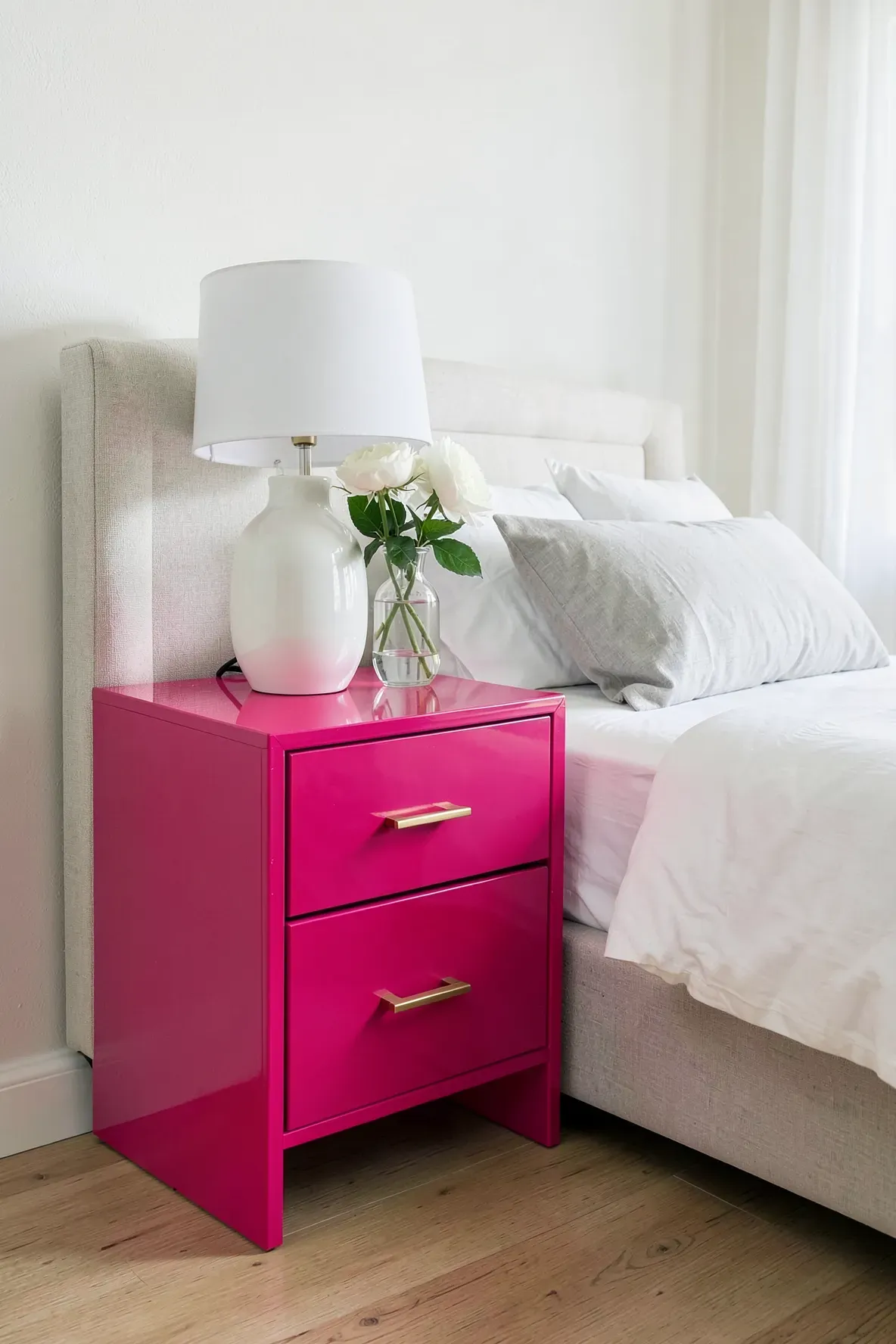 A glossy hot pink nightstand with brass handles next to a bed with white linens.