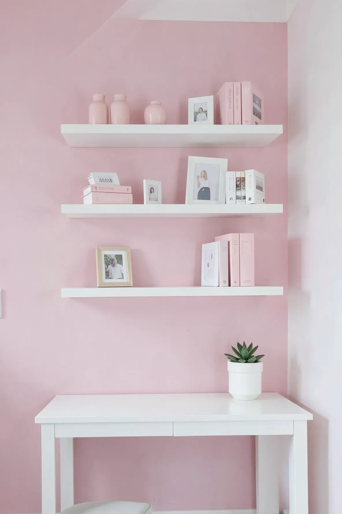 White floating shelves on a pink wall holding organized books and pink decor.